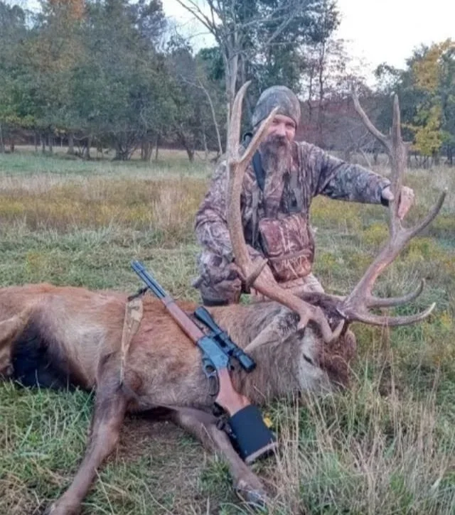 Hunter in camouflage gear kneeling next to a large dead deer with antlers, lying on the grass in a field during daytime.