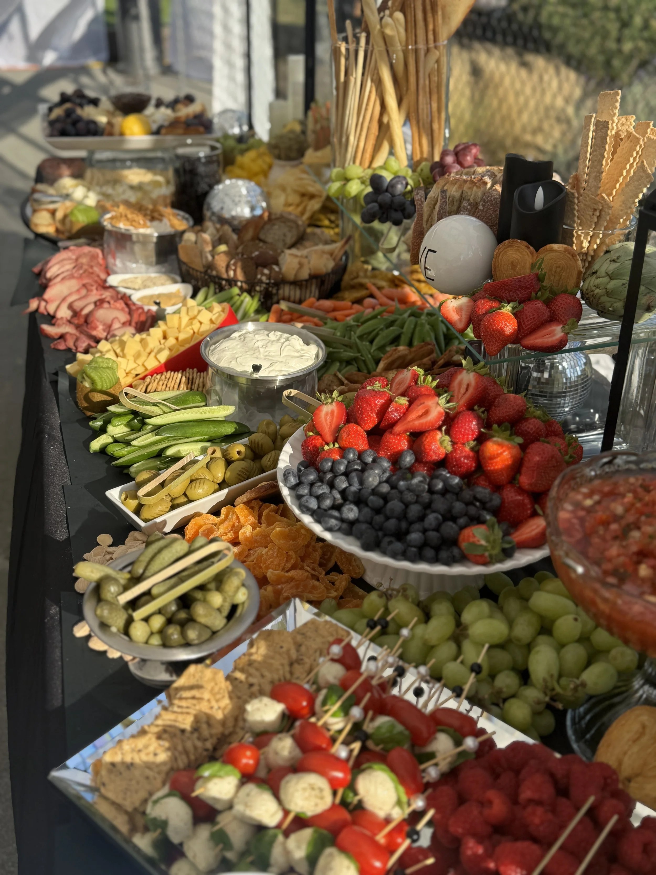 A buffet table filled with various appetizers and snacks, including fresh strawberries, blueberries, grapes, sliced vegetables, cheese, crackers, bread, and skewers of cherry tomatoes, mozzarella, and basil. There are also dips, breadsticks, and other finger foods arranged for a party or gathering.