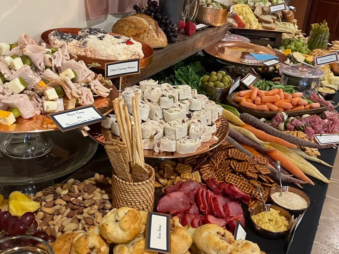 A large spread of various cheeses, meats, fruits, vegetables, nuts, crackers, and bread displayed on a buffet table with labeled dishes.