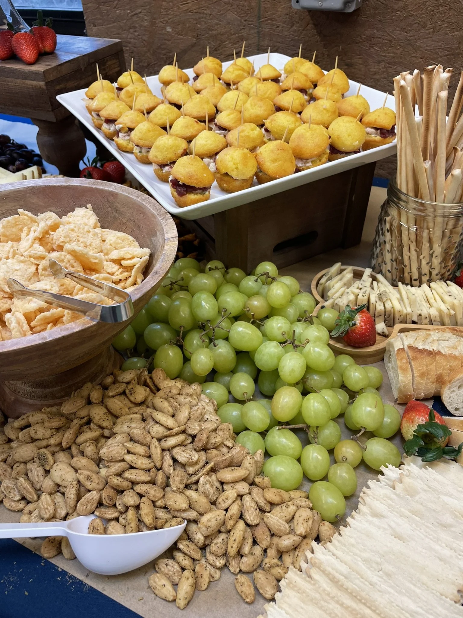 A display of various snacks and fruits, including small baked appetizer bites on a tray, a bowl of peanuts, green grapes, strawberries, and some cheese slices.