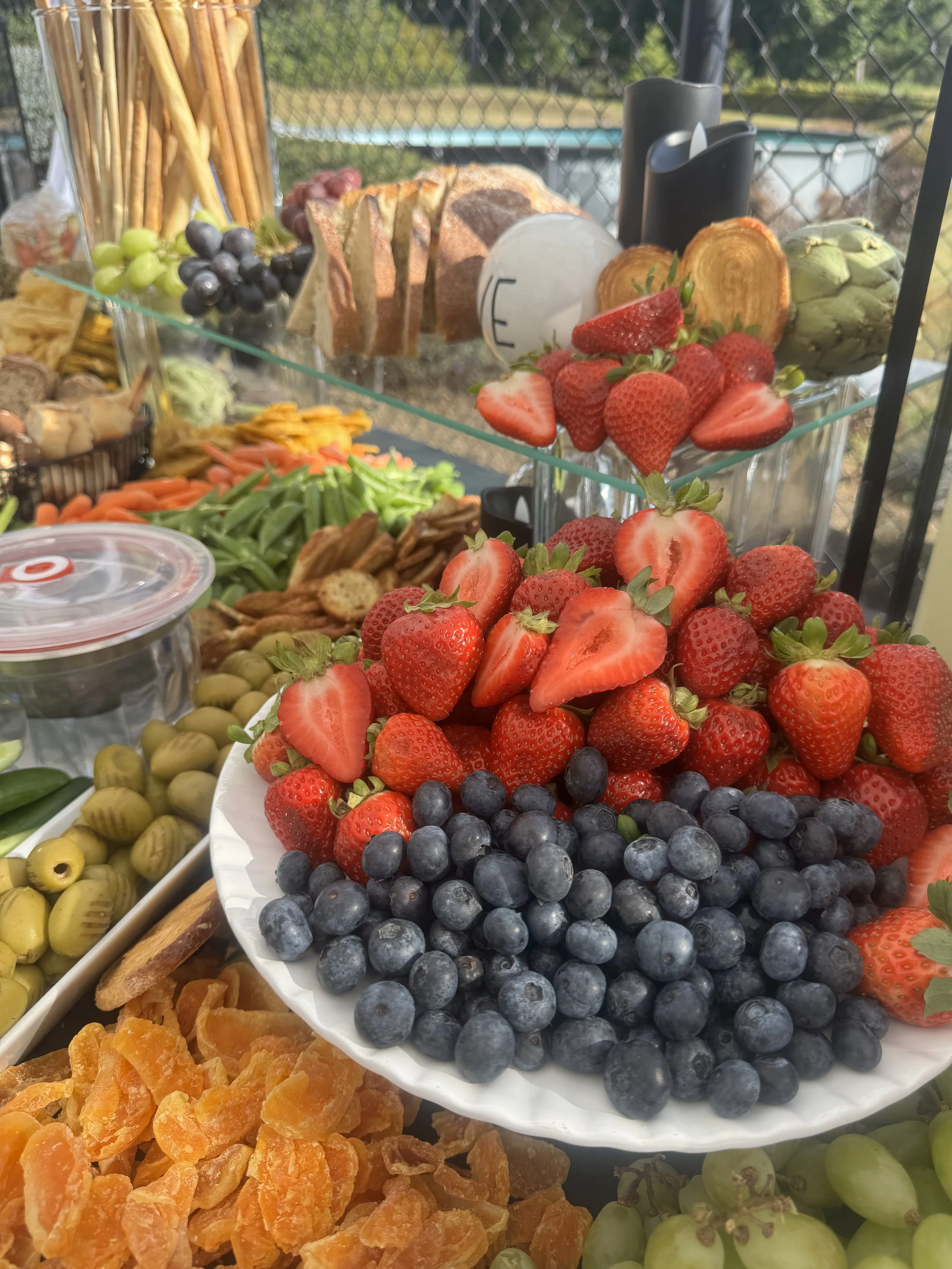 A display of fresh strawberries, blueberries, and artichokes on a white plate, with other assorted fruits and snacks on a table at an outdoor setting.