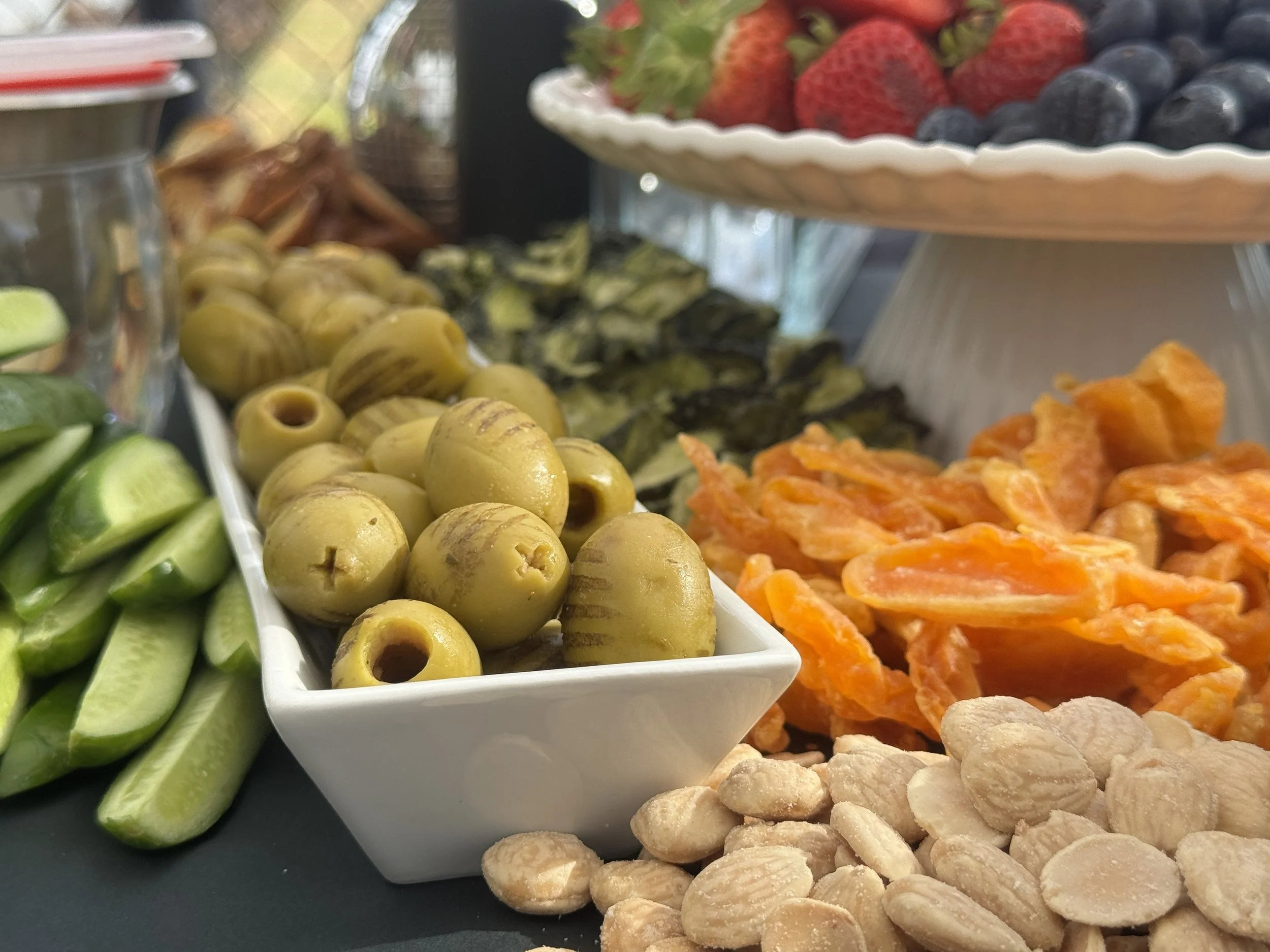 Assorted fresh fruits and snacks displayed on a black surface, including grapes, strawberries, blueberries, pickles, and crackers in small bowls.