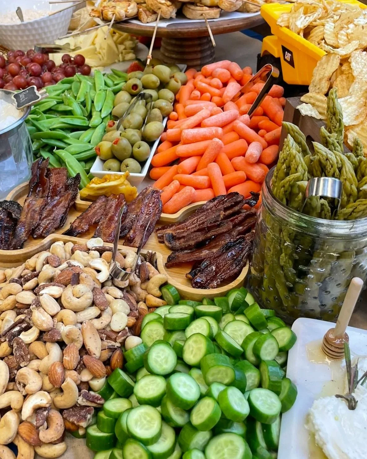 Assorted fresh vegetables and snacks on a buffet table, including carrots, cucumbers, green beans, olives, nuts, bacon, and chips.