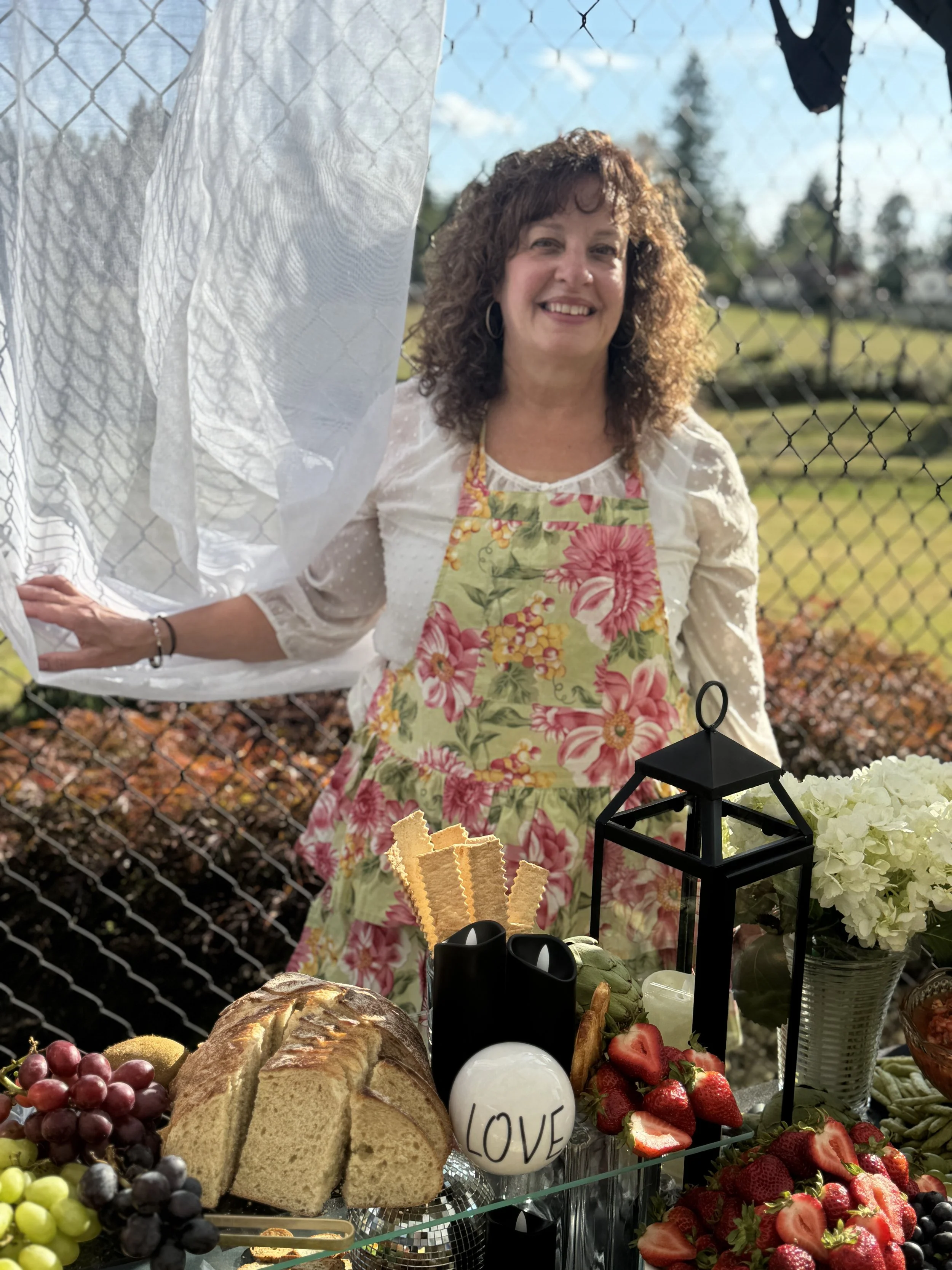 Woman smiling behind a fence during an outdoor gathering, with food and flowers on a table in the foreground.