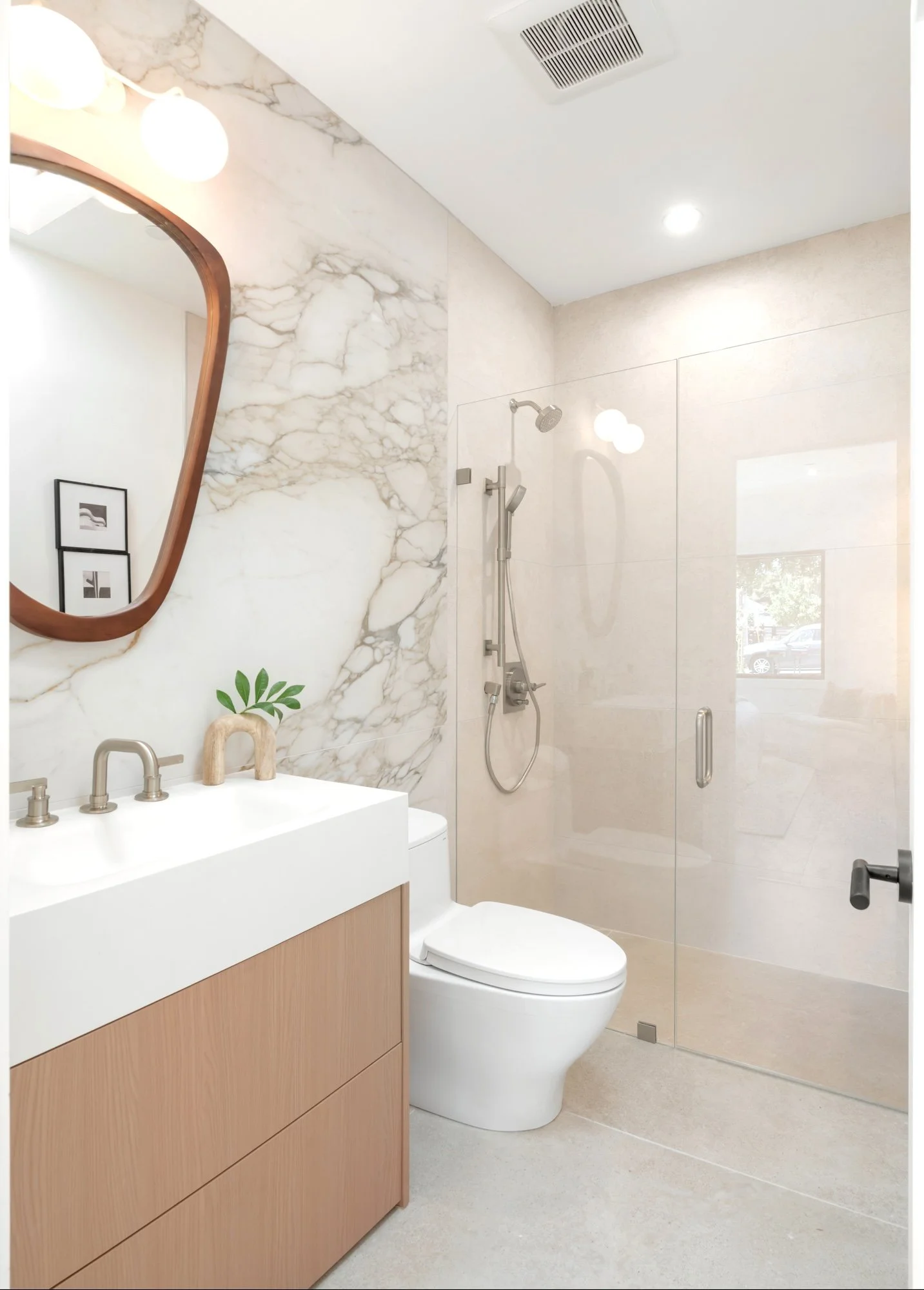 Modern bathroom with a marble wall, mirror, wooden vanity, and a toilet. Shower with glass doors and a rain showerhead, small window, and minimal decoration.