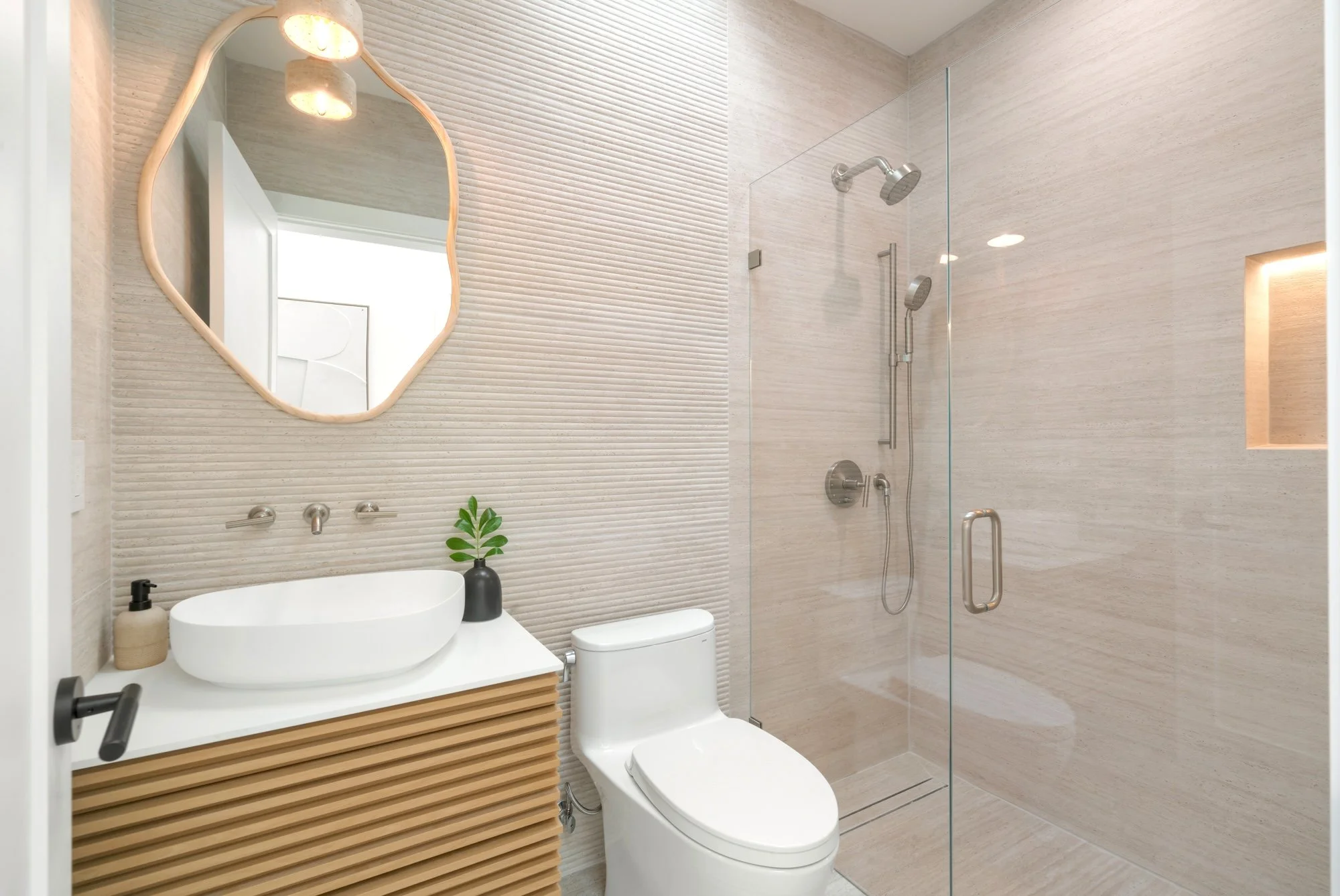 Modern bathroom with textured beige walls, a white vessel sink on a wooden vanity, a beige soap dispenser and a potted plant. There is a mirror with a scalloped wooden frame, a wall-mounted faucet, a toilet, and a walk-in shower with a glass door, a 