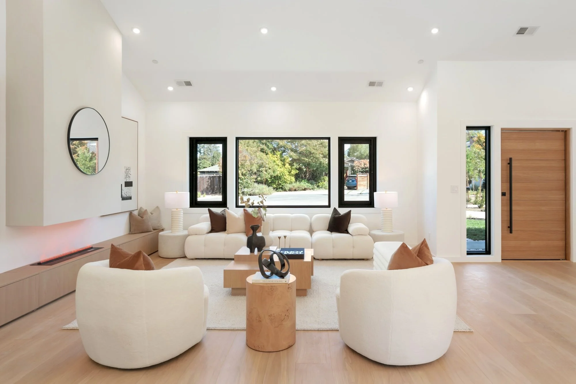 Bright living room with white walls and ceiling, large windows with black frames overlooking a green backyard, cream upholstered sofas, brown and beige throw pillows, modern wooden side tables, decorative items, and minimalistic design.