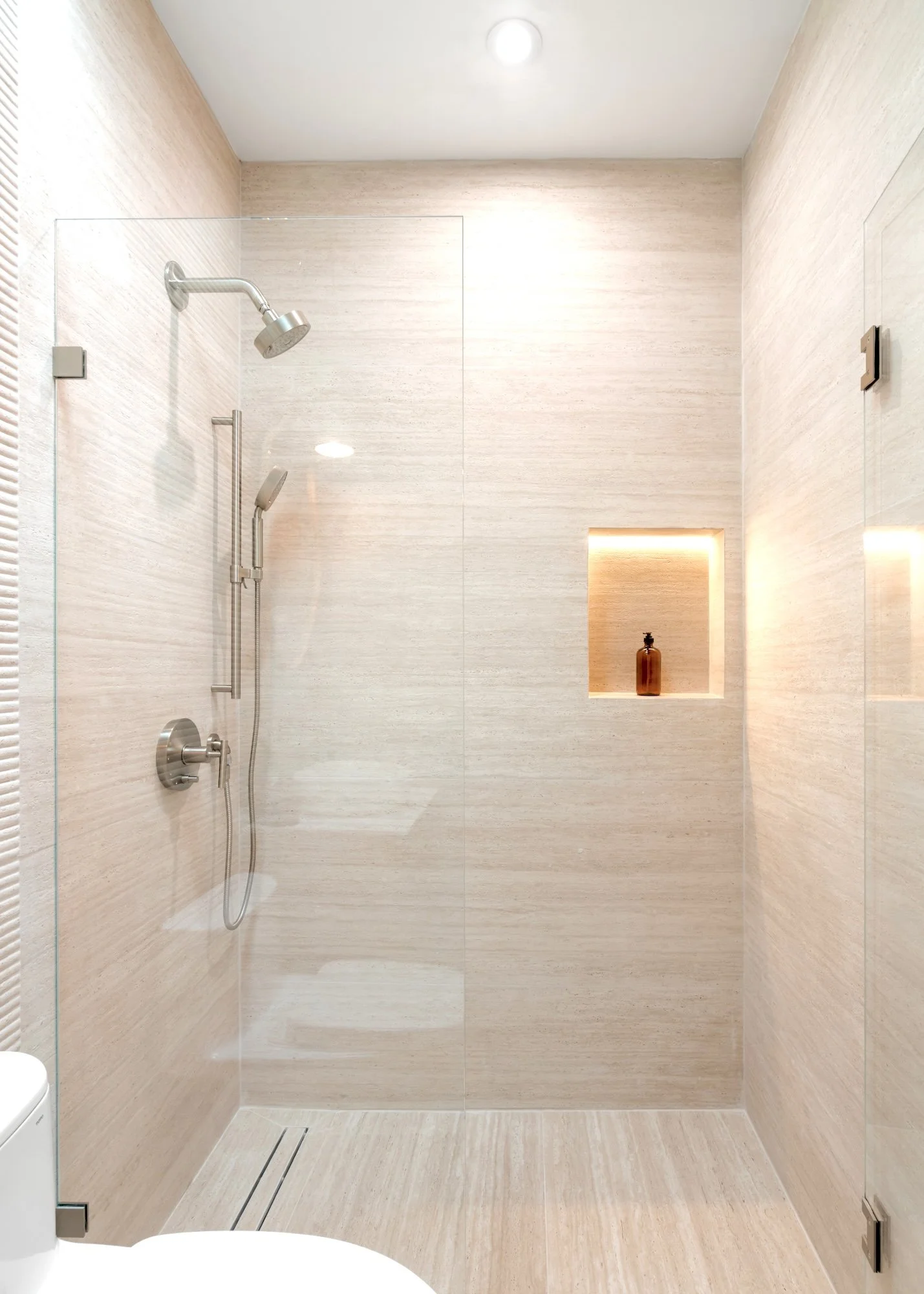 Modern walk-in shower with glass door, beige tile walls and floor, overhead and handheld showerheads, built-in niche with a brown bottle, and a ceiling light.
