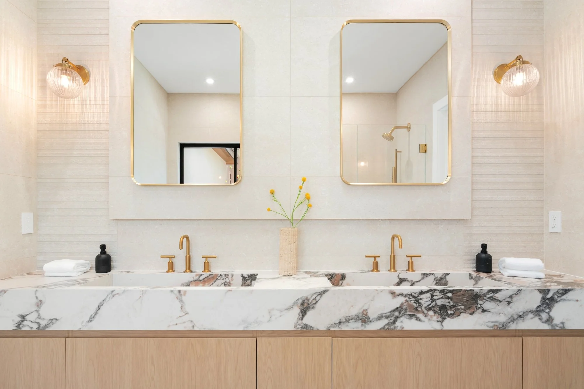 Bathroom vanity with two mirrors, gold fixtures, black soap dispensers, and a vase with yellow flowers.
