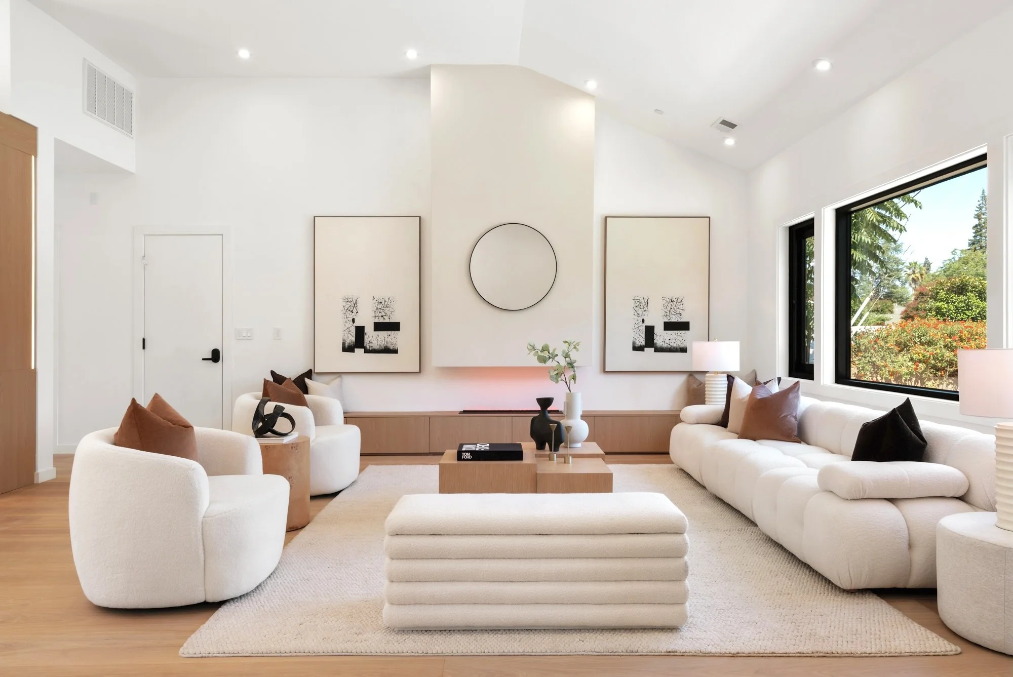 Modern living room with white walls, large windows showing greenery outside, contemporary art on the walls, white sofas with brown and black pillows, a wooden coffee table, and a white textured rug.