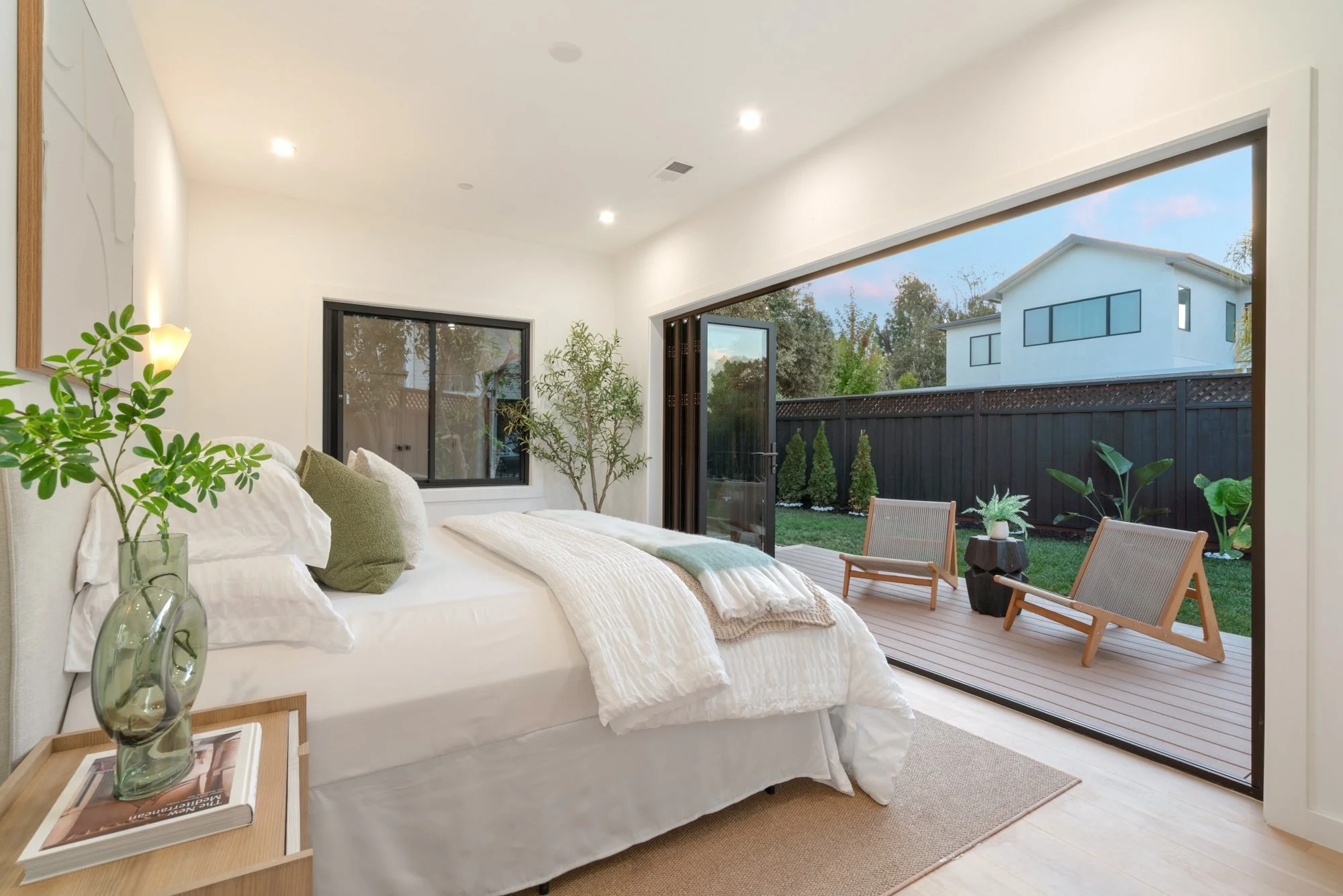 Bright bedroom with large sliding glass door opening to a backyard patio with chairs and small plants. Inside, there is a bed with white linens, green pillows, and a side table with vases and magazines. The room has white walls and ceiling, with rece