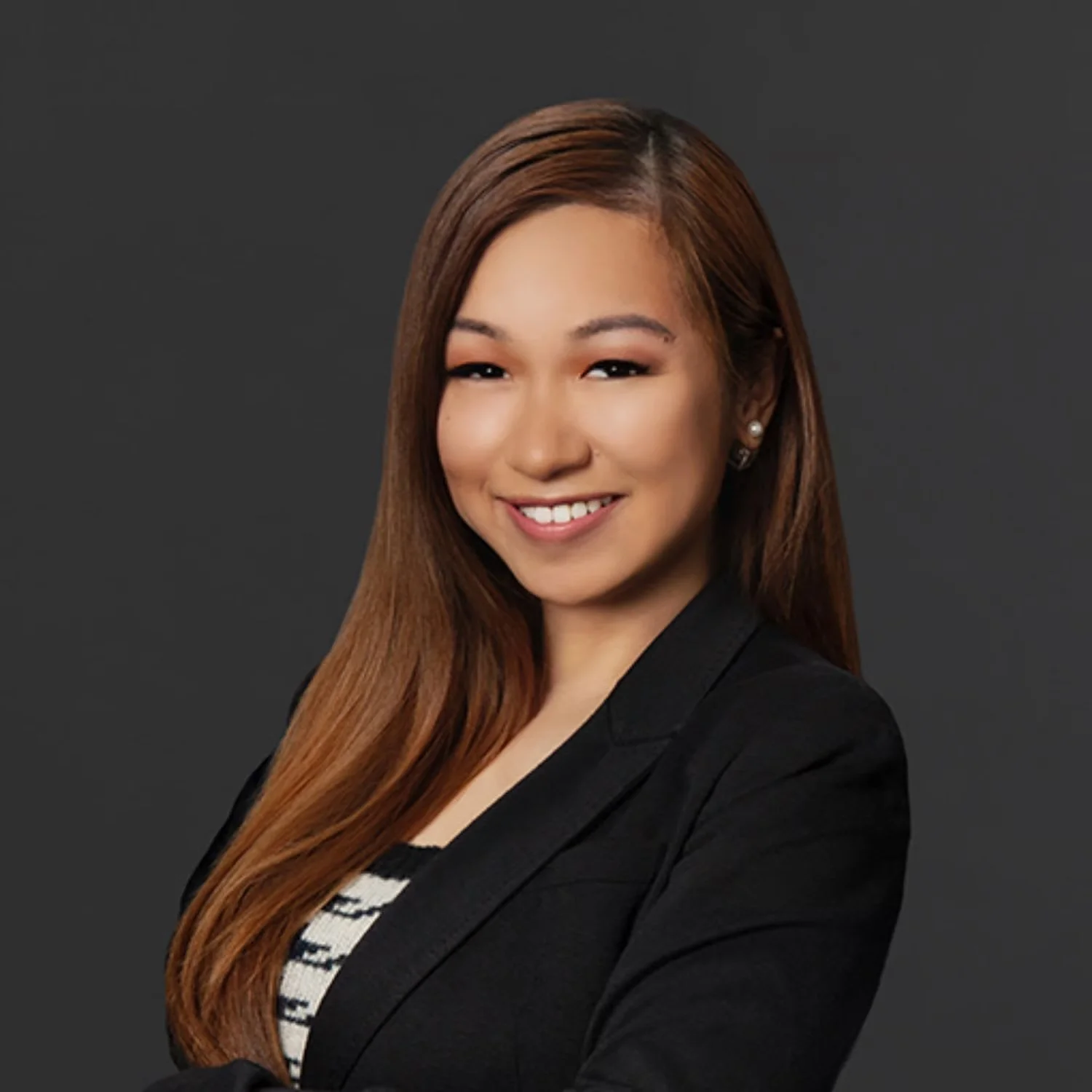 Professional portrait of a smiling woman with long brown hair, wearing a black blazer and a striped top, against a dark gray background.