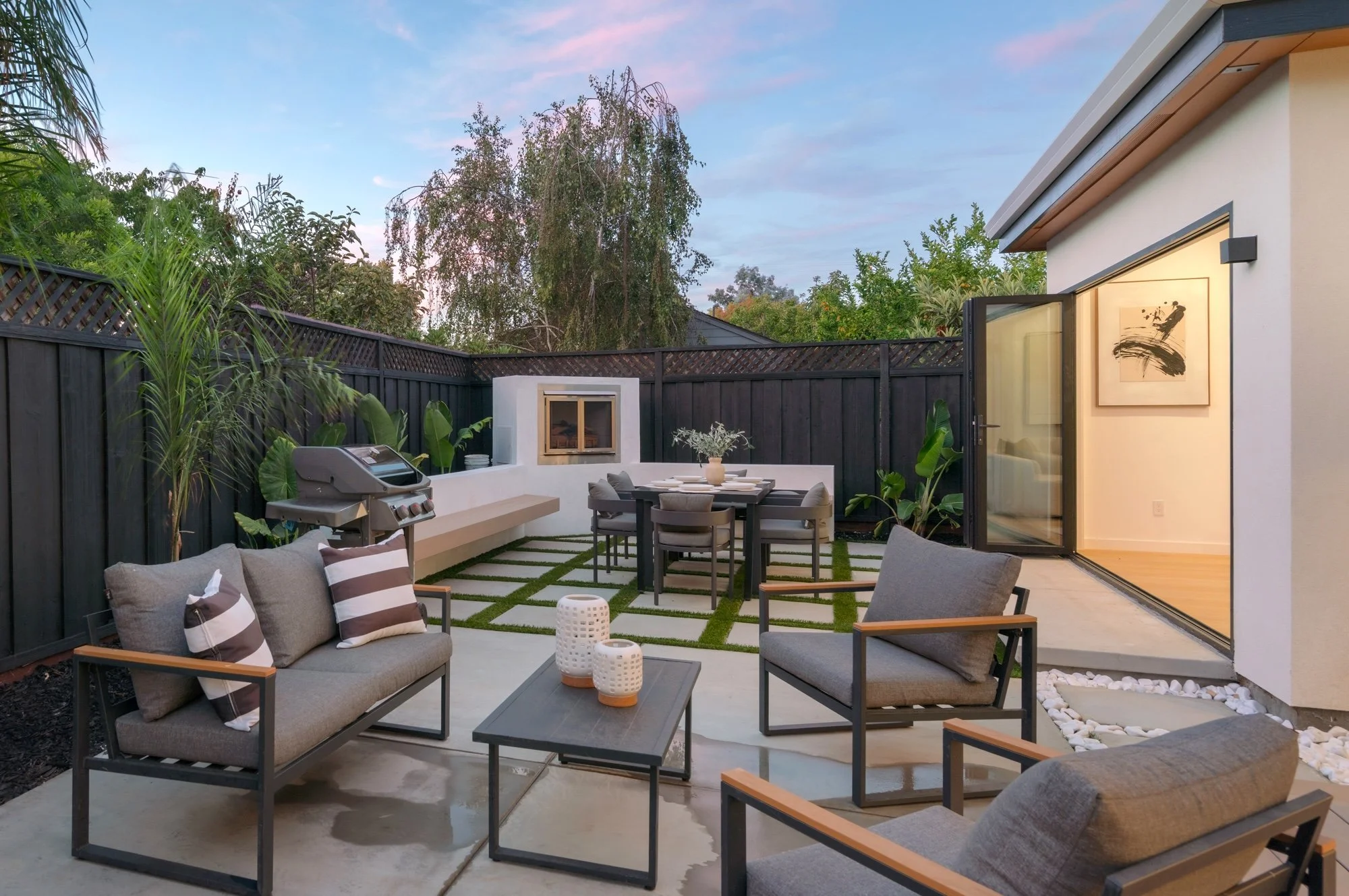 Modern backyard patio with seating area, outdoor dining table, barbecue grill, and decorative plants, enclosed by a black fencing and adjacent to a house with sliding glass doors.