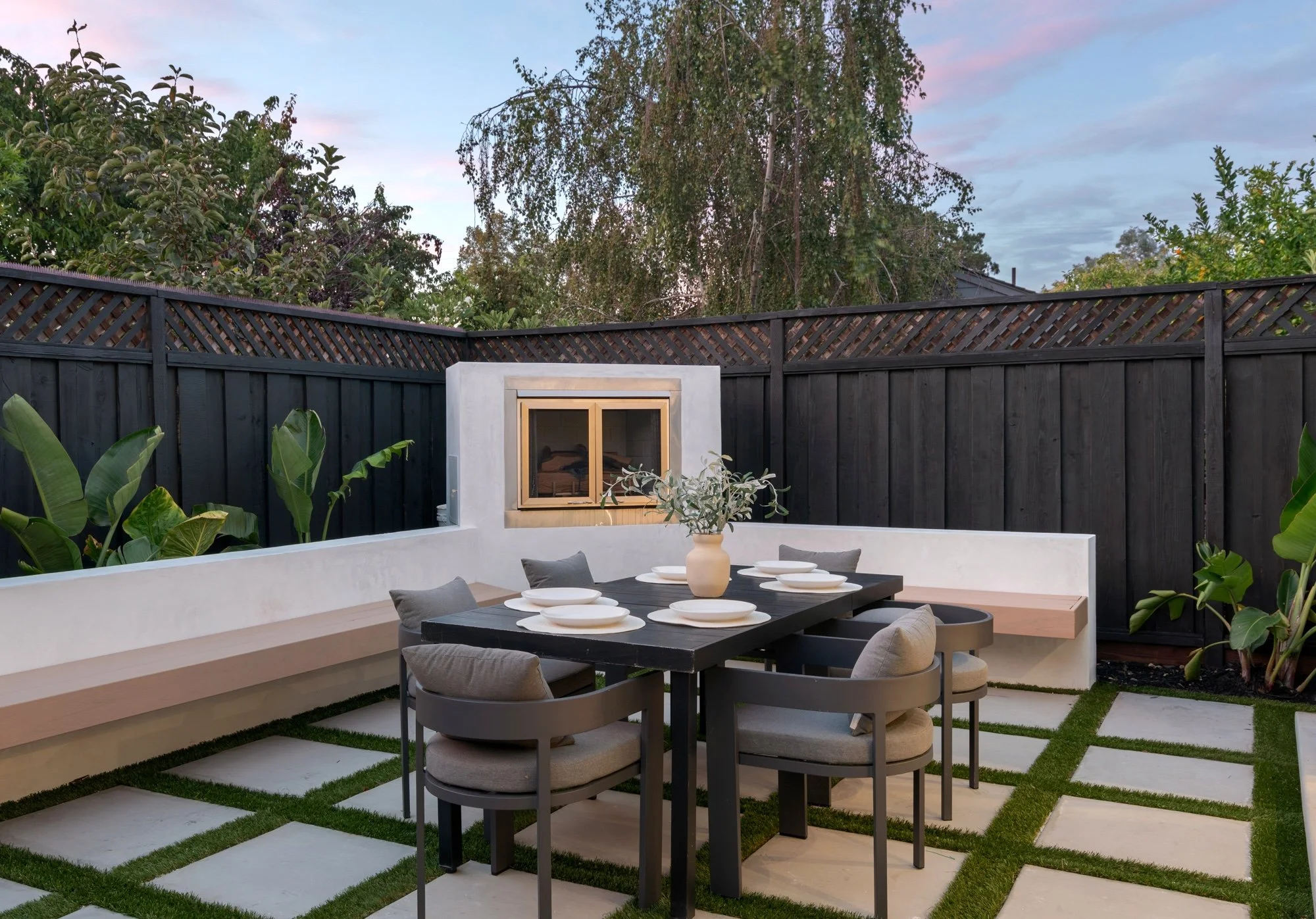 An outdoor dining area with a rectangular table set with white plates, a beige vase, and surrounded by six cushioned chairs, enclosed by a black wooden fence and plants, during dusk.