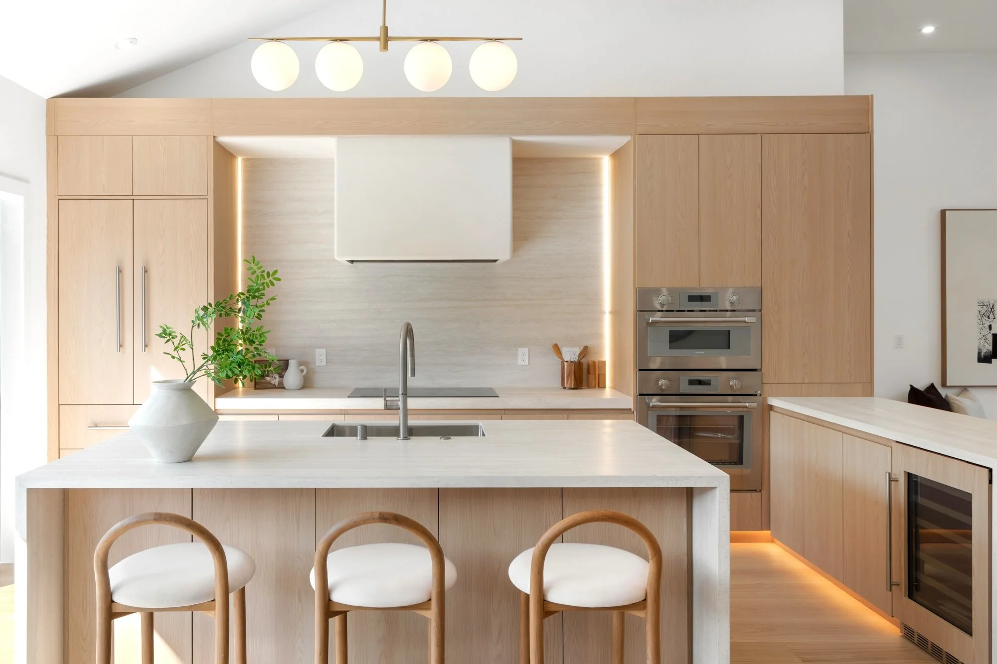 Modern kitchen with light wood cabinetry, white countertops, a central island with three white chairs, and built-in stainless steel appliances. A potted plant and decorative items are on the counter, illuminated with soft lighting and a contemporary 