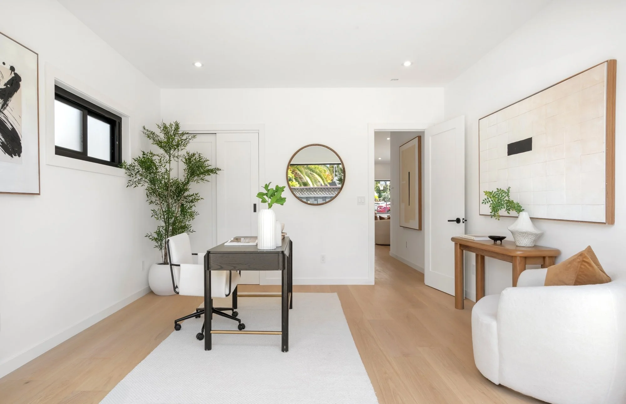 Modern living room with white walls, wooden flooring, a round mirror, and various art pieces. Contains a dining table with white chairs, a potted plant, a side table with a white vase, and a cozy white armchair with brown cushions.
