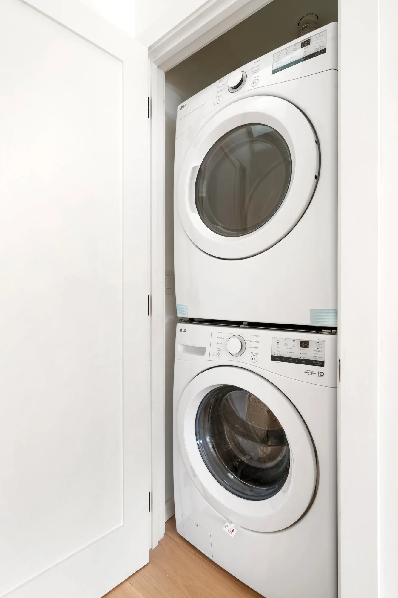 Stacked white washing machine and dryer in a small laundry closet with white walls and light wooden floor.