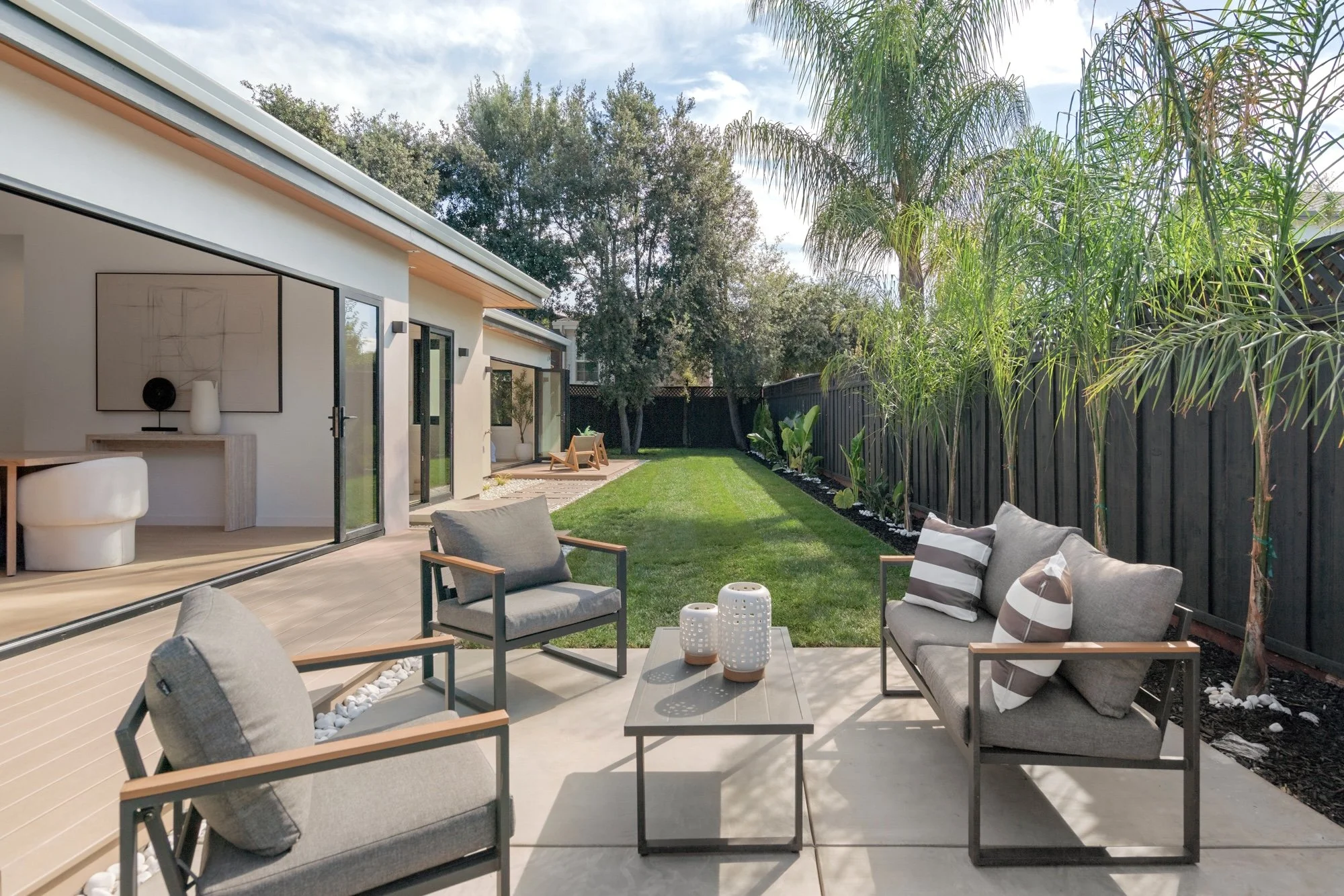 Outdoor backyard patio with modern outdoor furniture, including gray cushioned chairs, a sofa with striped pillows, and matching tables, surrounded by a lush green lawn, small decorative white rocks, and tall trees with a black fence in the backgroun
