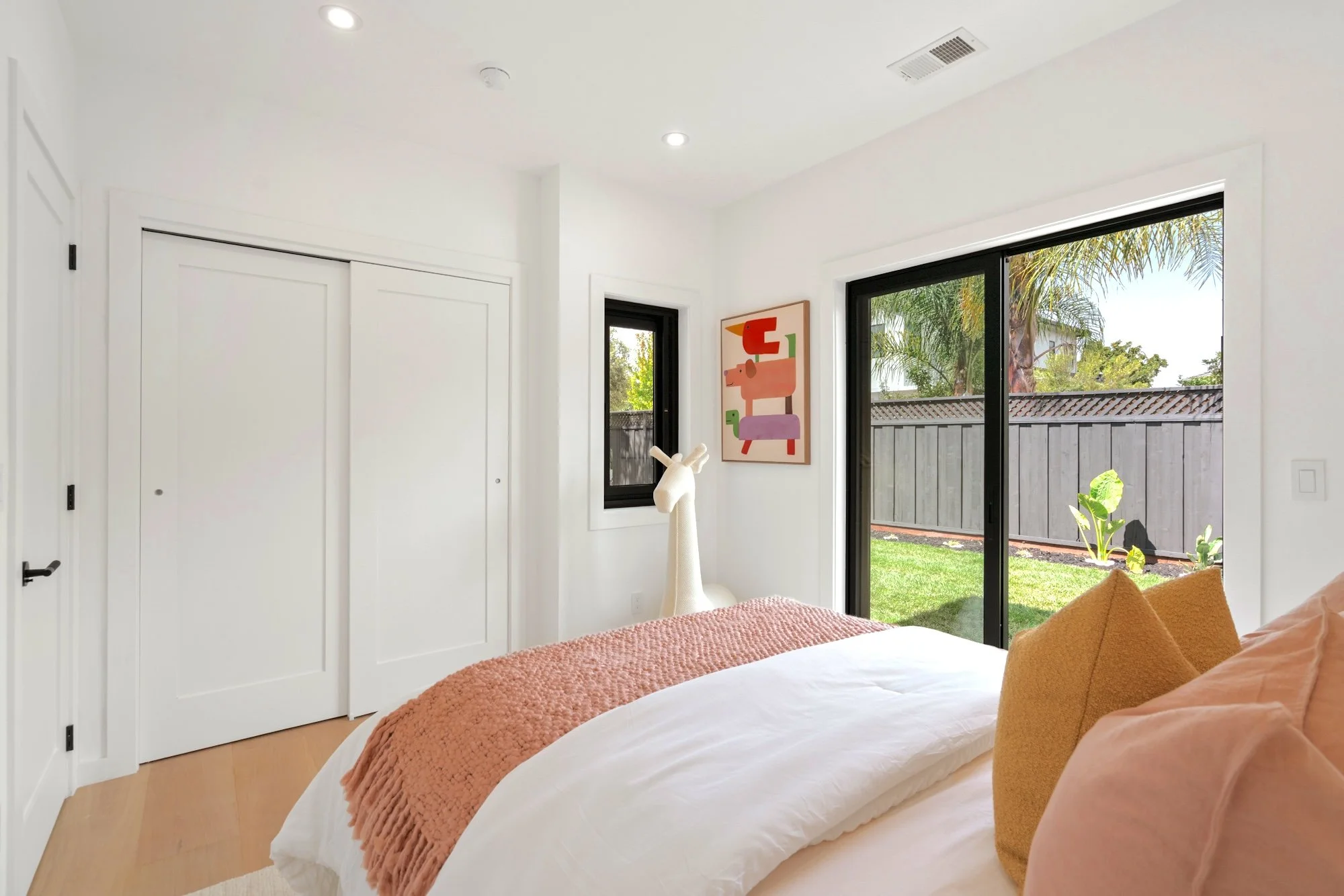 A bright bedroom with white walls and ceiling, featuring a large sliding glass door opening to a backyard with palm trees and a fence. The bed has white sheets, a peach-colored blanket, and matching pillows. A colorful framed animal art hangs on the 
