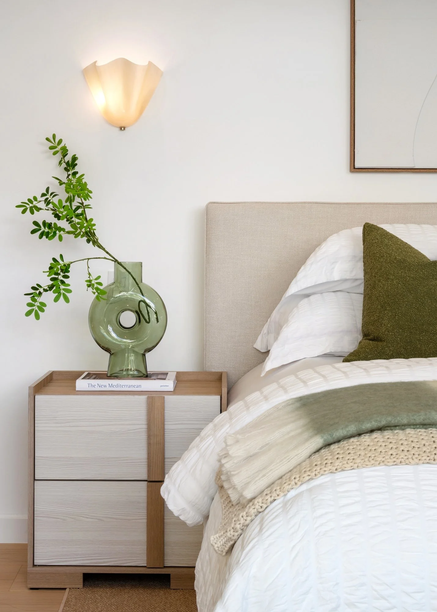 A bedroom corner with a nightstand, a vase with green foliage, a bed with white and green bedding, a wall lamp, and a framed art piece on a white wall.