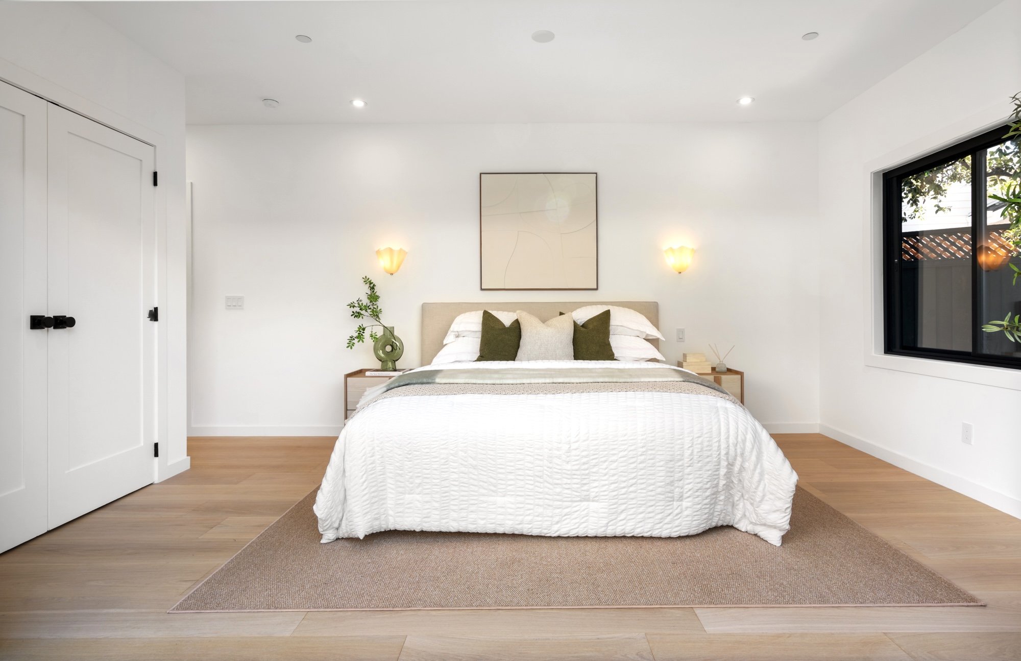 A minimalistic bedroom with white walls, a white bed with textured bedding, green and beige pillows, a wooden nightstand with a book and a green vase with a plant, wall-mounted lamps, a large abstract artwork above the bed, a window with black trim s