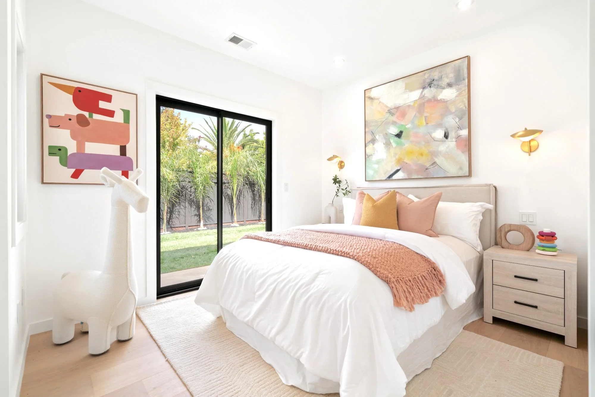 A bright, modern bedroom with white walls, a large bed with white bedding and peach-colored pillows, abstract paintings on the walls, a wooden nightstand with a small stack of colorful items, a white deer-shaped sculpture, a sliding glass door reveal