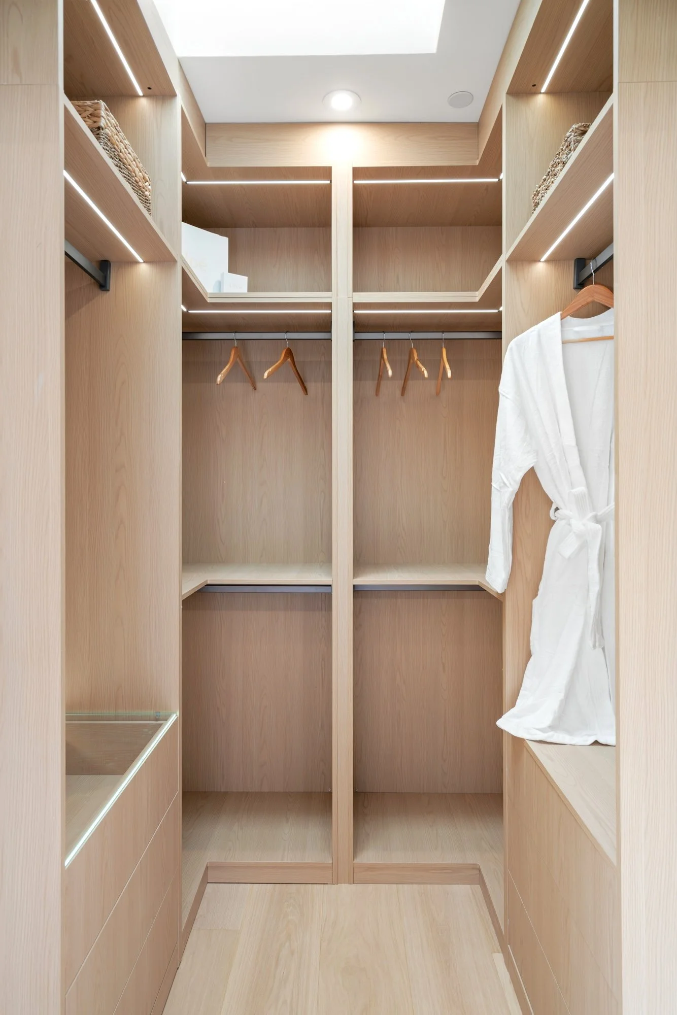 Empty walk-in closet with light wood shelves, hanging rod with hangers, and a white bathrobe hanging on the right side. Built-in LED lighting and a skylight overhead.