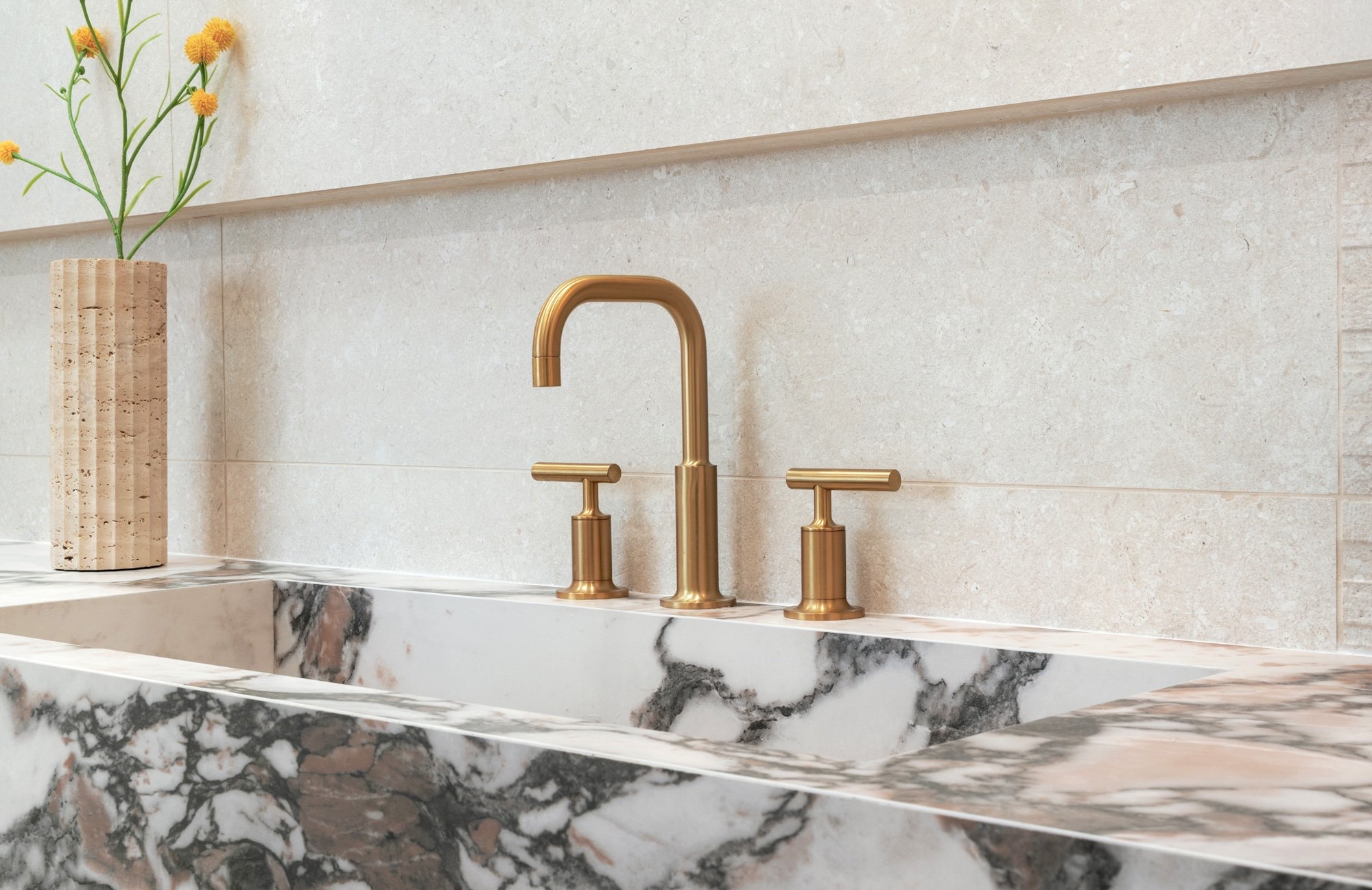 Close-up of a modern bathroom sink with a marble countertop in black, white, and brown tones, a shiny gold faucet with two handles, and a beige textured wall with a decorative trim. There is a tall, rectangular vase with vertical grooves holding a gr