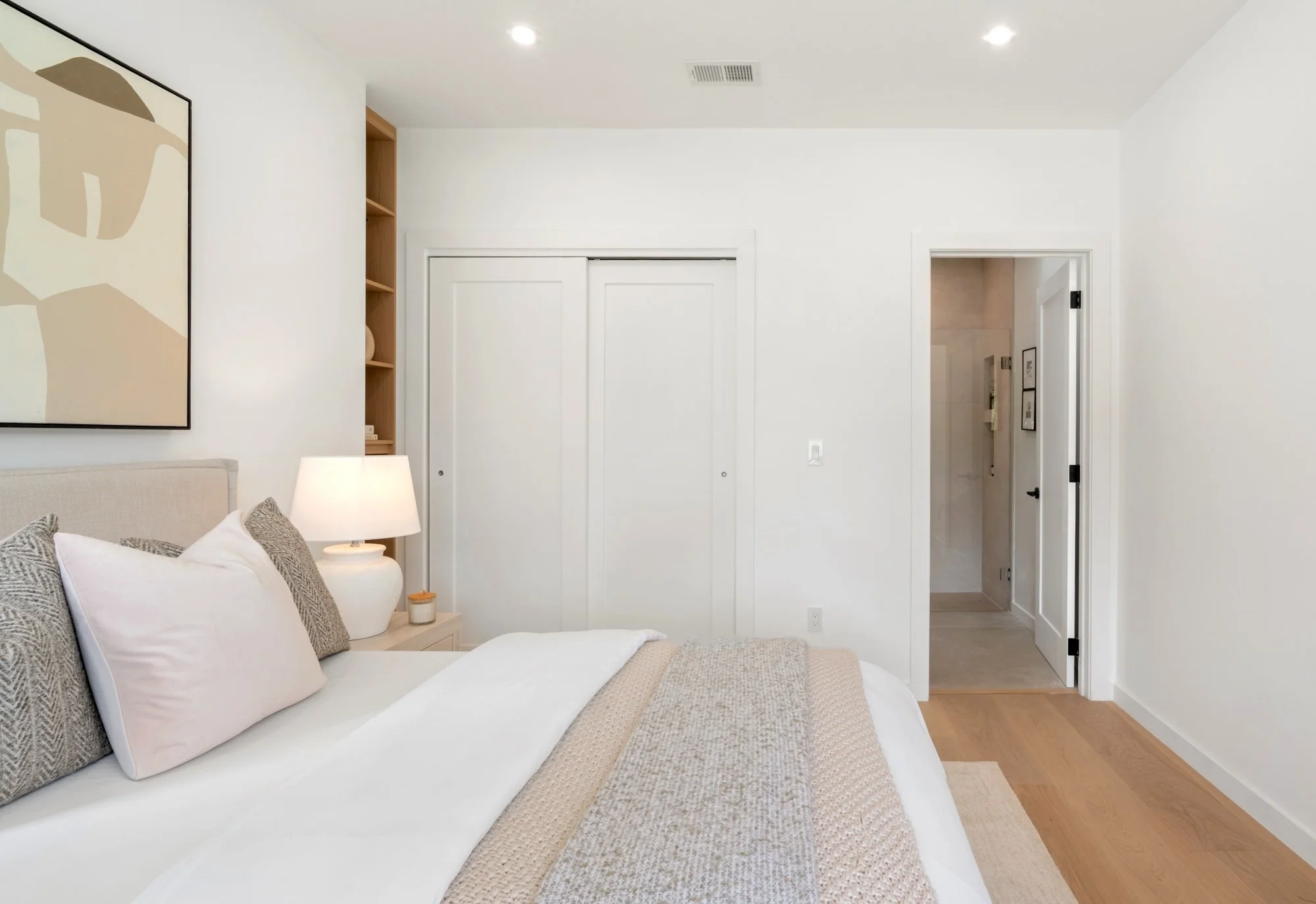A bedroom with a bed, pillows, a nightstand, a lamp, and a painting on the wall. There is a closet with sliding doors and an open door leading to a bathroom.