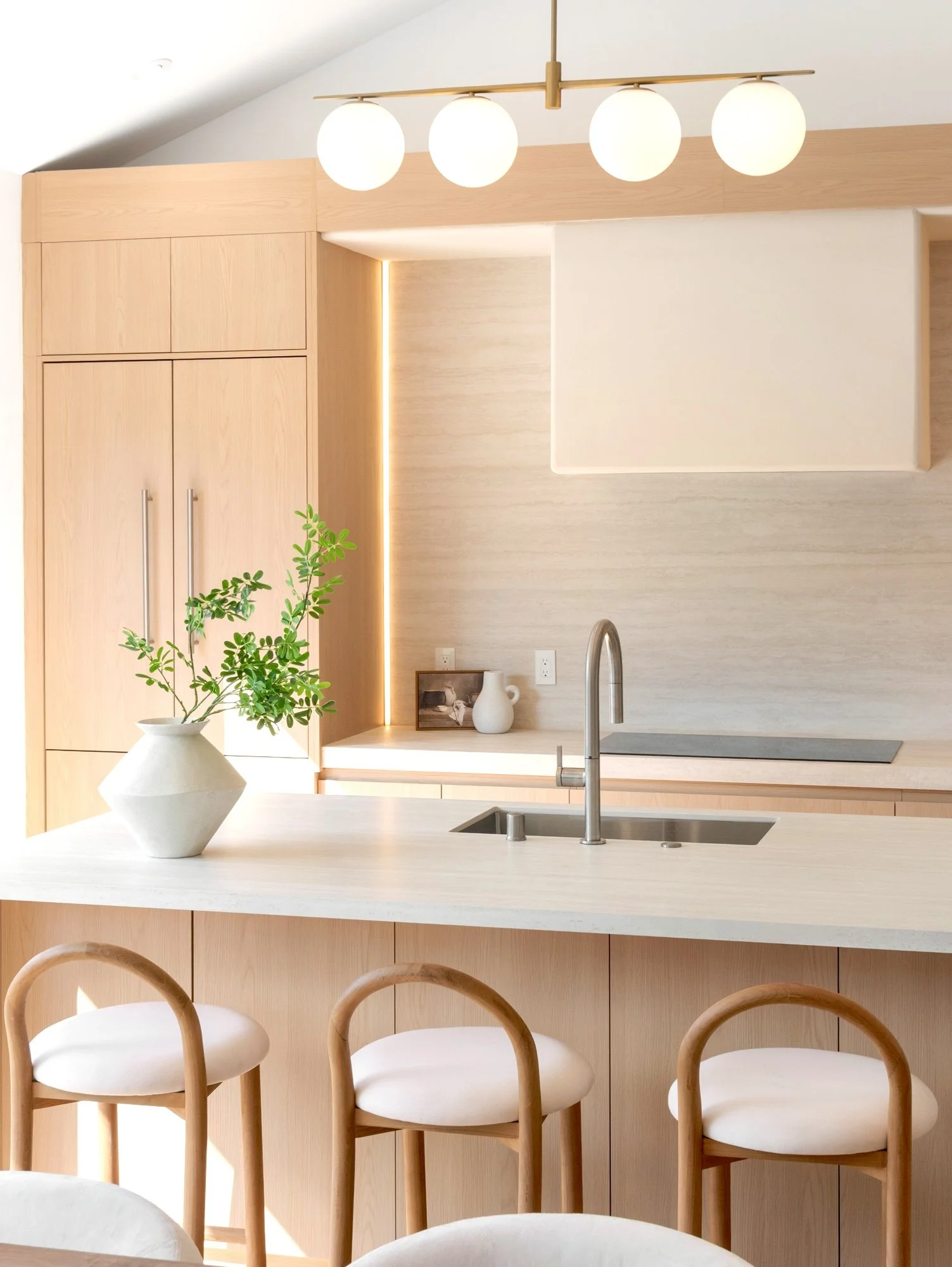 A modern kitchen with a white countertop, a stainless steel faucet, light wood cabinets, and a white vase with a green plant on the counter. There are four chairs with curved wooden backs and white seats, and a wooden light fixture with four round bu