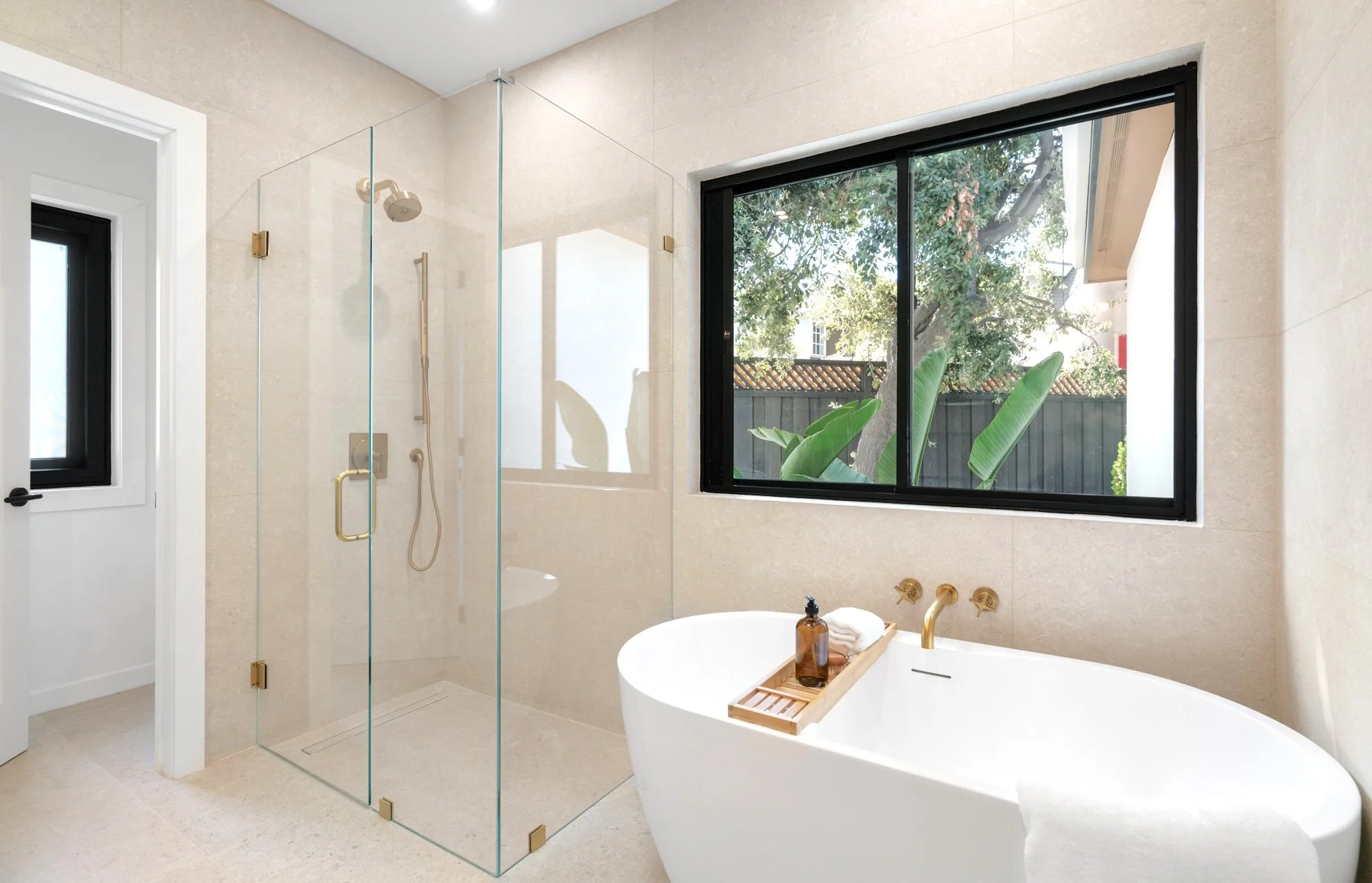 Modern bathroom with a large window, beige tile walls, a glass shower enclosure with gold fixtures, and a freestanding bathtub with a wooden tray holding toiletries.