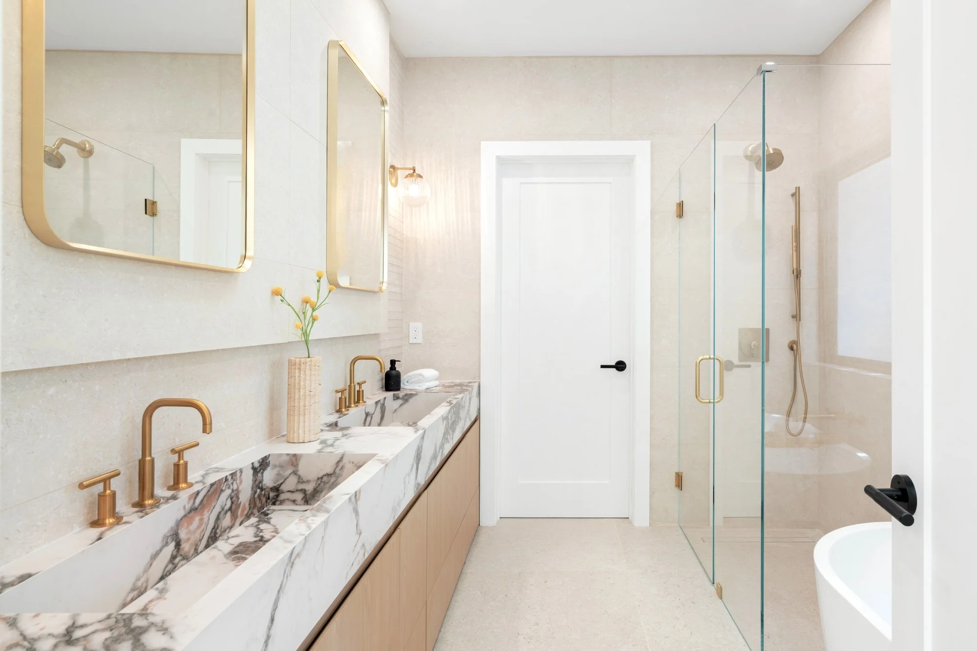 Modern bathroom with marble countertop vanity, gold fixtures, large mirrors, and a glass-enclosed shower with beige tiles.