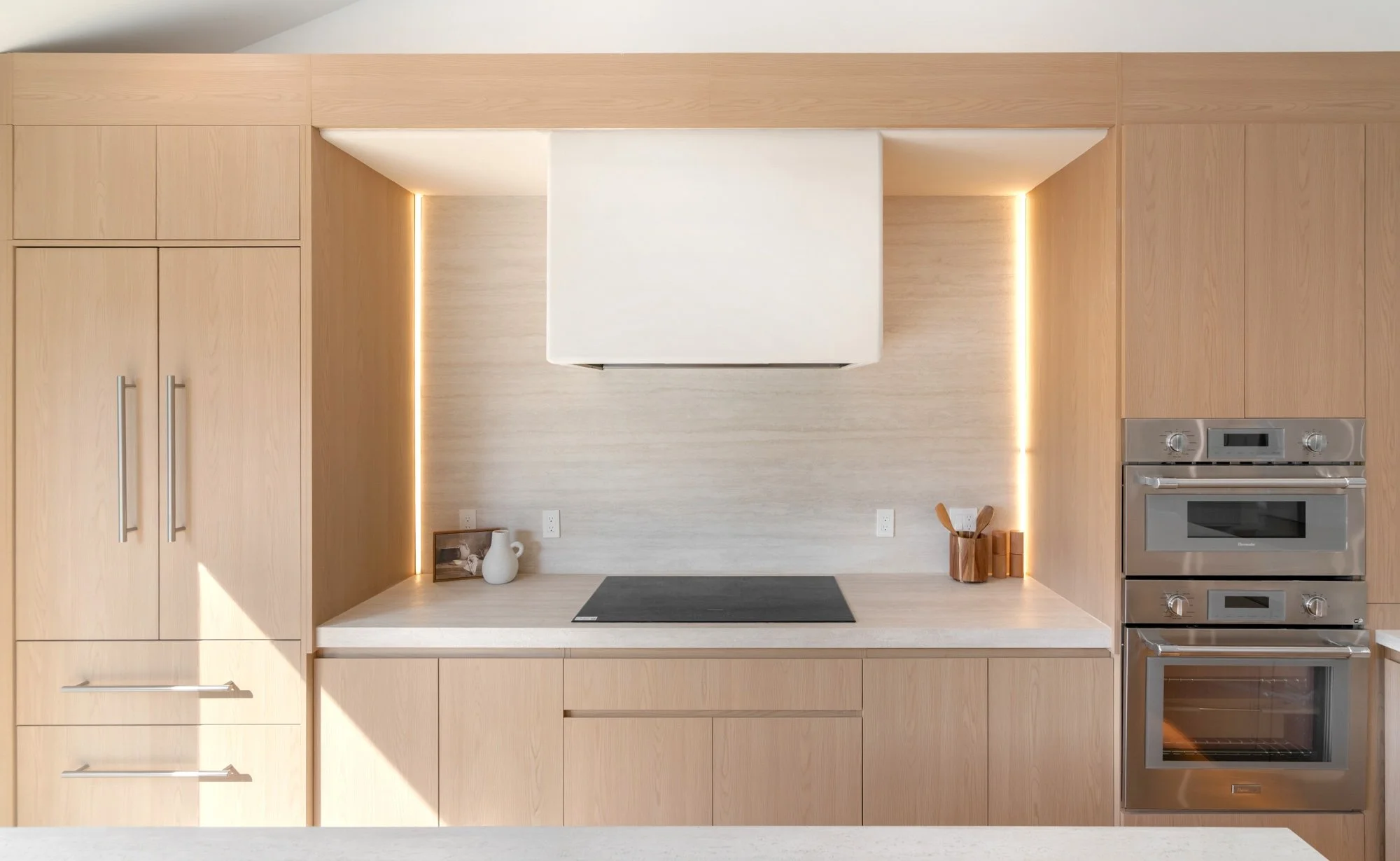 Modern kitchen with light wood cabinetry, built-in oven, white range hood, decorative vases, and minimal decor.