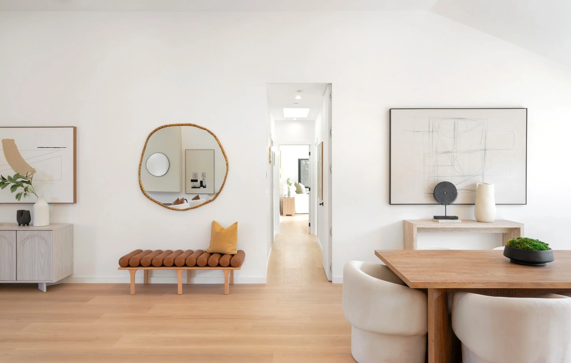 A modern, minimalist living room with white walls, abstract artwork, a small bench with a yellow pillow, a side table with a white vase and black decor, a round mirror, and a wooden dining table with white upholstered chairs.