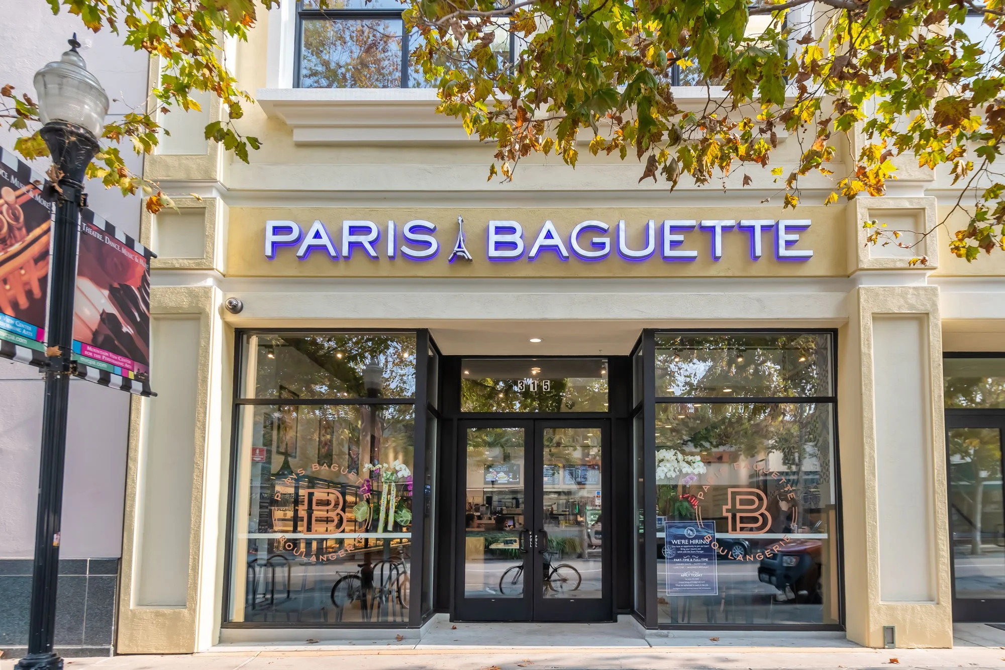 Front entrance of a Paris Baguette bakery and cafe with glass doors, an illuminated sign, a sidewalk, and trees with fall-colored leaves overhead.