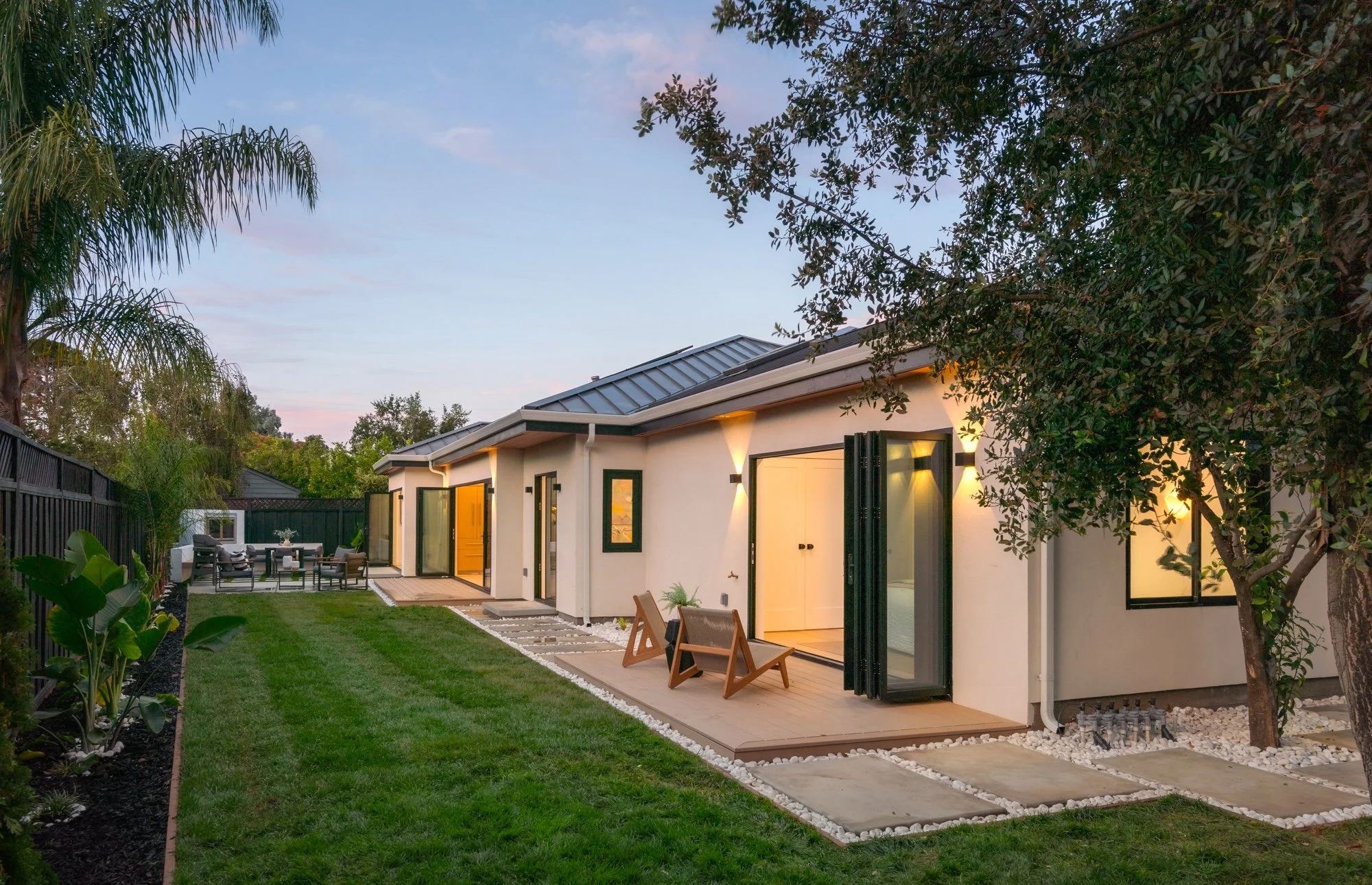 A backyard scene during dusk with a modern white house featuring black-framed windows and doors, surrounded by lush greenery including a tall palm tree and other trees, a fenced yard with outdoor furniture, and a well-maintained lawn and pathway.