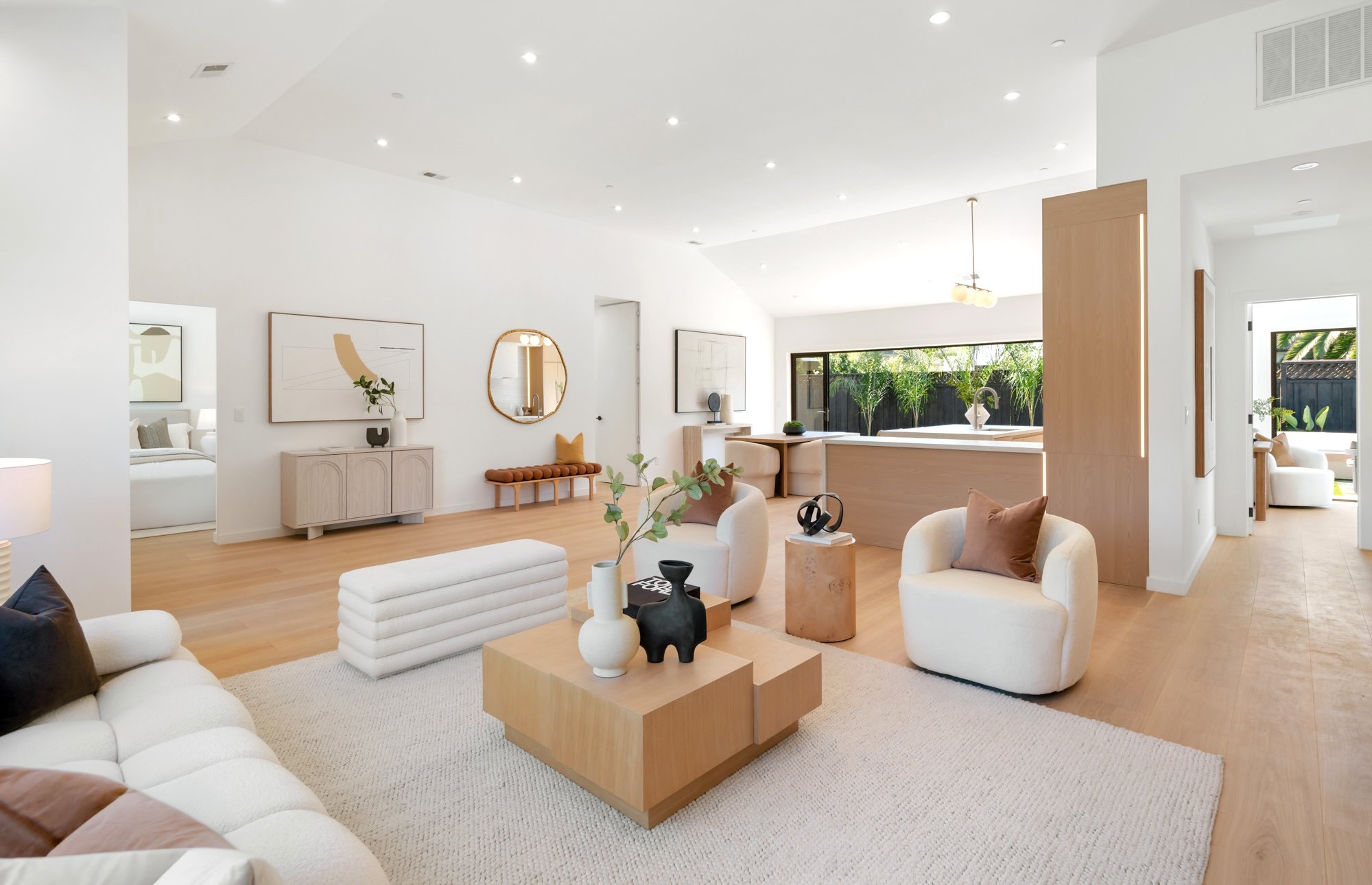 Bright and airy living room with white walls, light wood flooring, modern white and beige furniture, and minimalist decor. Large windows and sliding glass door open to a backyard with greenery.