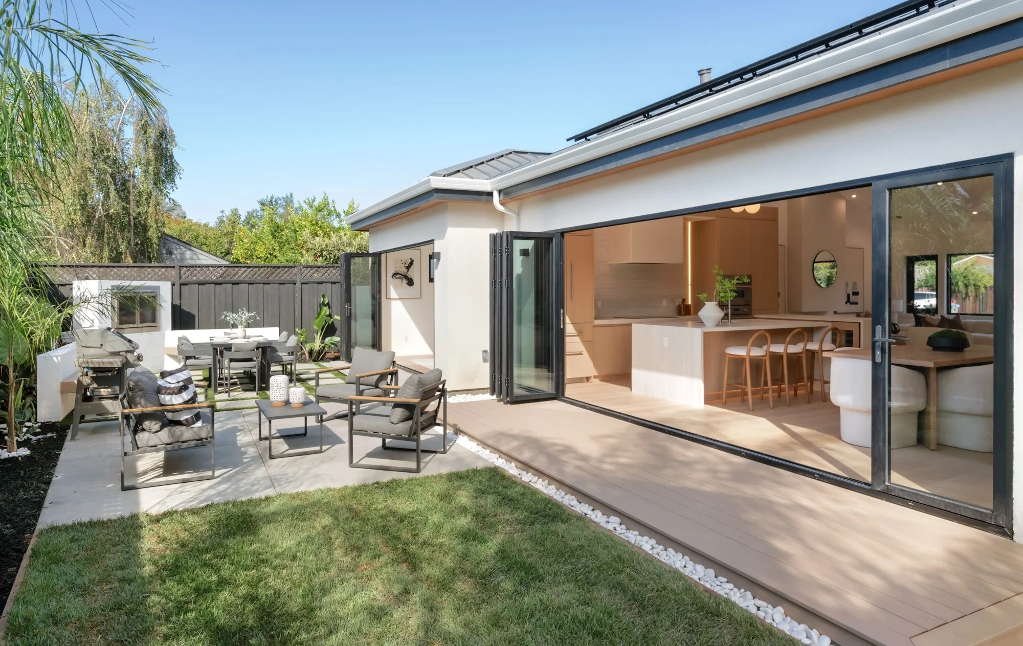 Backyard patio with outdoor seating, plants, and a view into a modern interior kitchen and living area via large glass doors.
