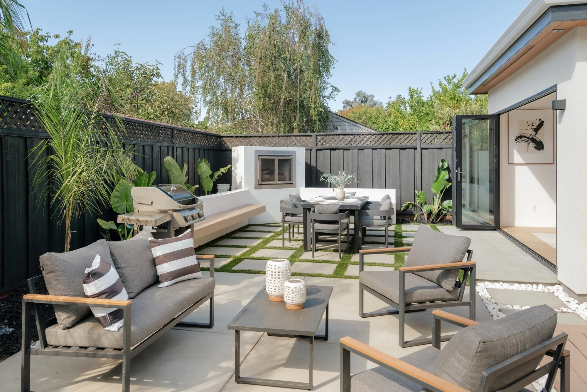 A modern outdoor patio area with gray seating, a dining table with surrounding chairs, a barbecue grill, and decorative plants enclosed by a black wooden fence, with a clear blue sky overhead.
