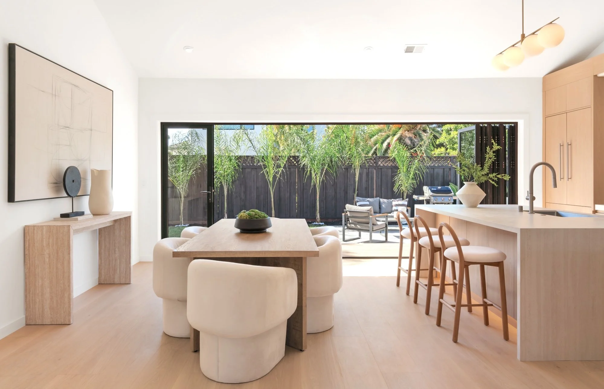 Modern dining and kitchen area with a view of an outdoor patio and garden, featuring trees and outdoor furniture.