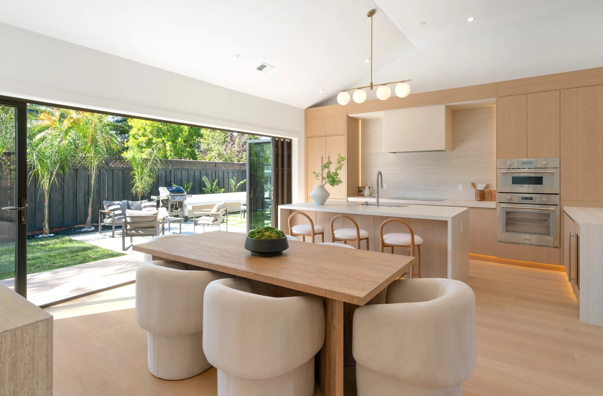 Modern kitchen and dining area with a view of an outdoor patio featuring seating and greenery, large sliding glass doors, and minimalist decor.