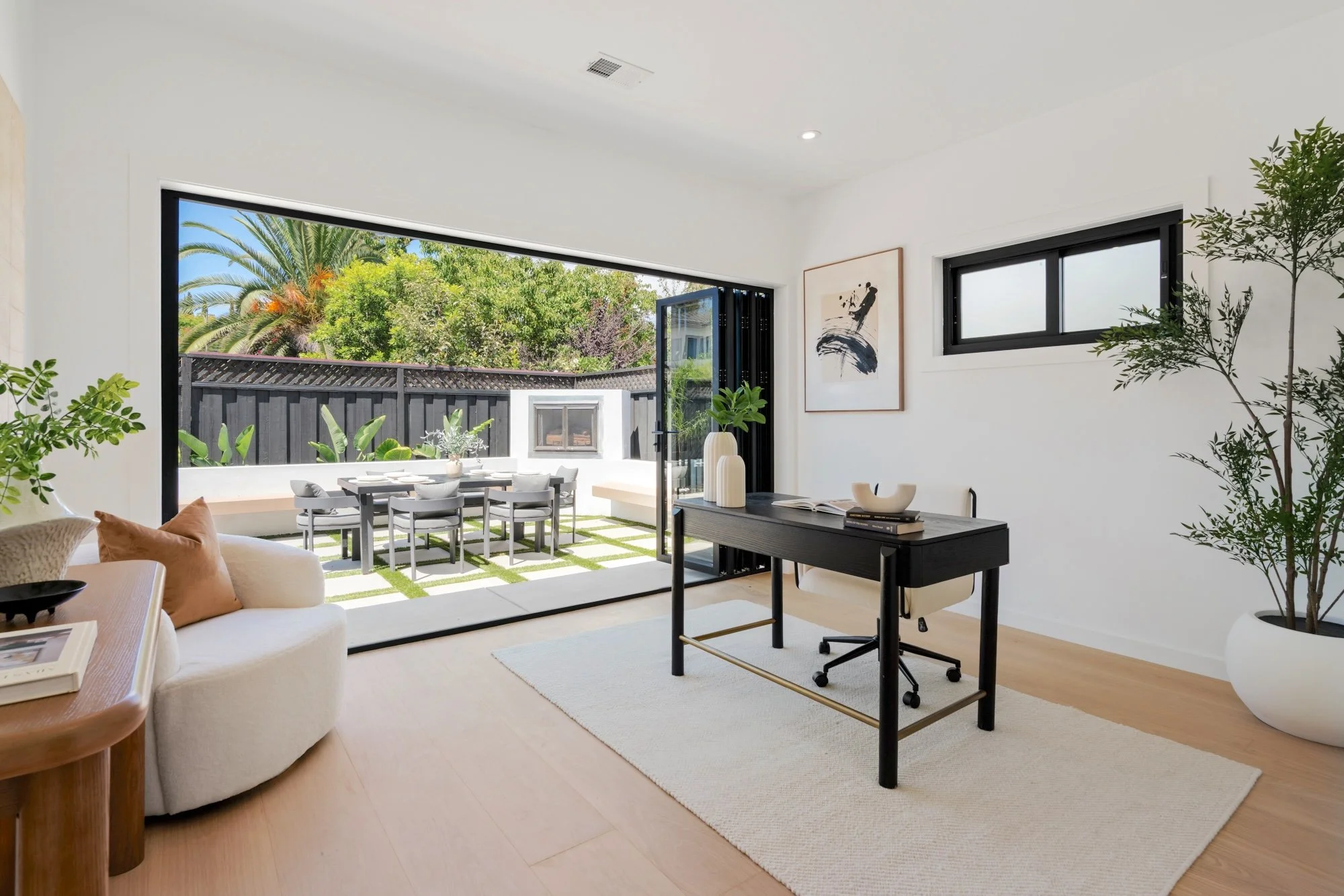 Bright living room with open sliding glass door leading to outdoor patio with dining table and chairs, lush greenery, and palm trees.