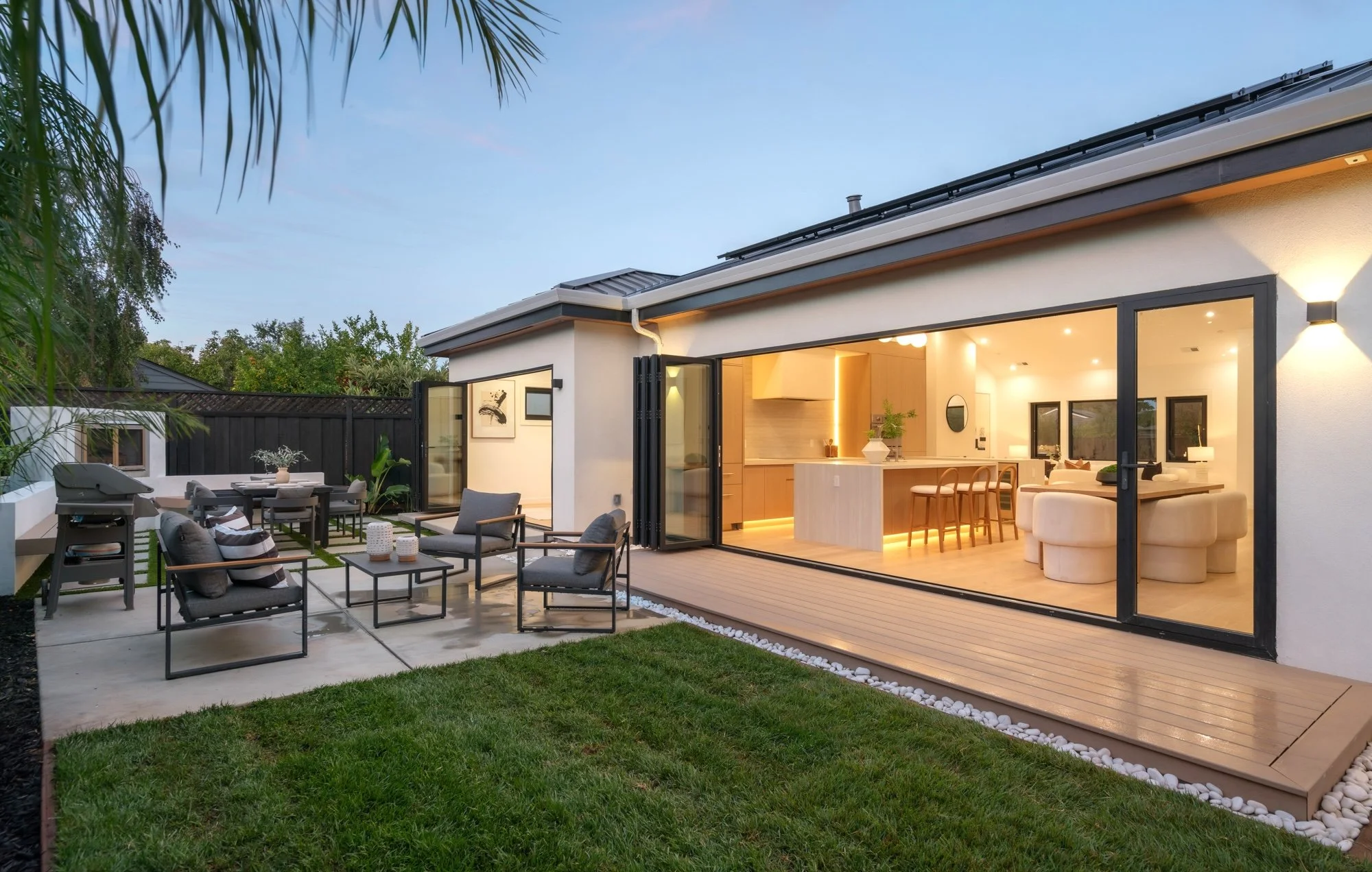 Modern backyard patio with outdoor seating, umbrella, grill, and an open sliding glass door revealing a contemporary indoor living space with kitchen and seating area, surrounded by a black fence and green trees.