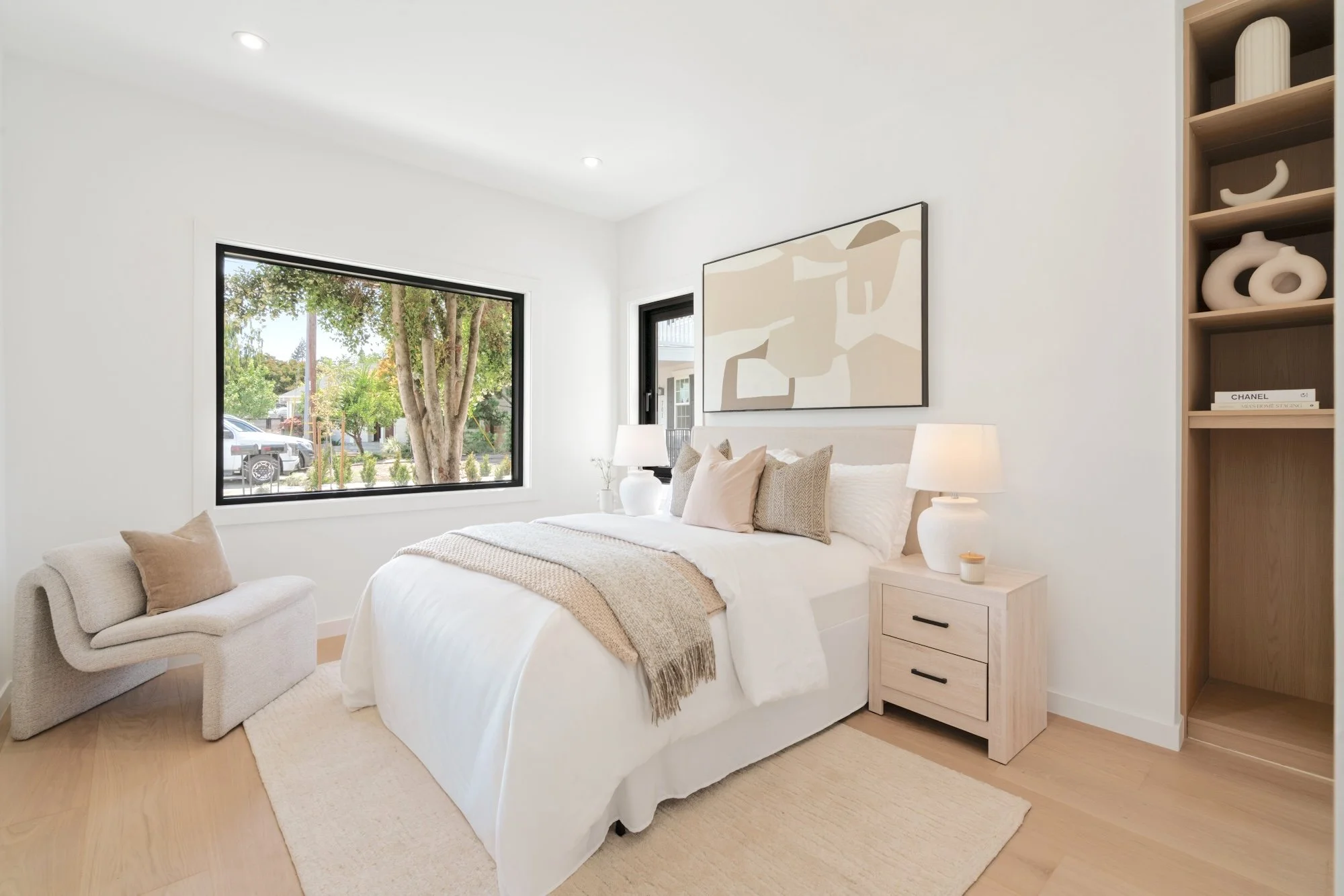 A bright modern bedroom with white walls and large windows showing trees and parked cars outside. The room features a white bed with beige and cream pillows, a beige throw blanket, and a small wooden nightstand with two drawers and black handles. Whi