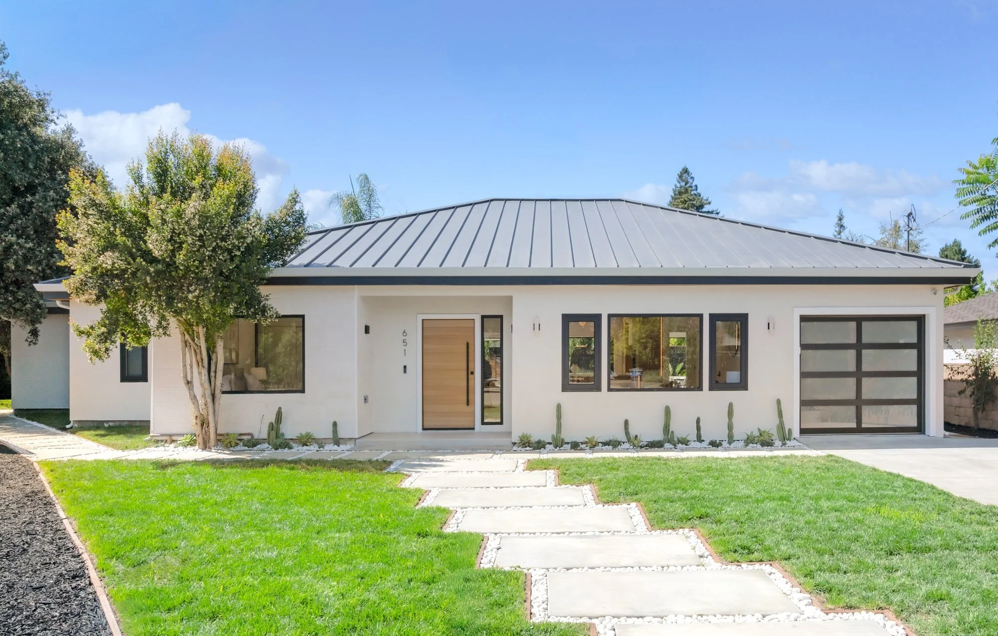 Front view of a modern single-story house with white exterior walls, a metal roof, large front windows, and a garage with a glass door, surrounded by a well-maintained lawn, trees, and a paved walkway.