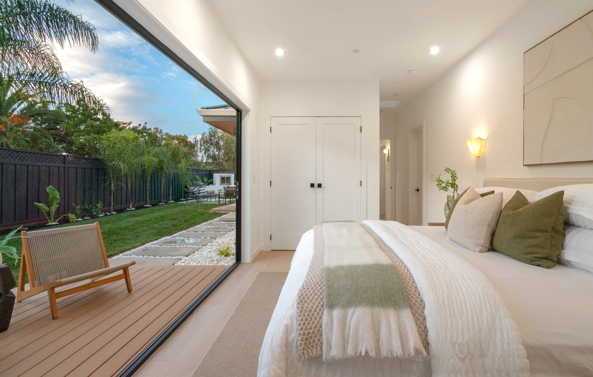Modern bedroom with large sliding glass door opening to a lush backyard with trees, outdoor furniture, and a stone paver pathway.
