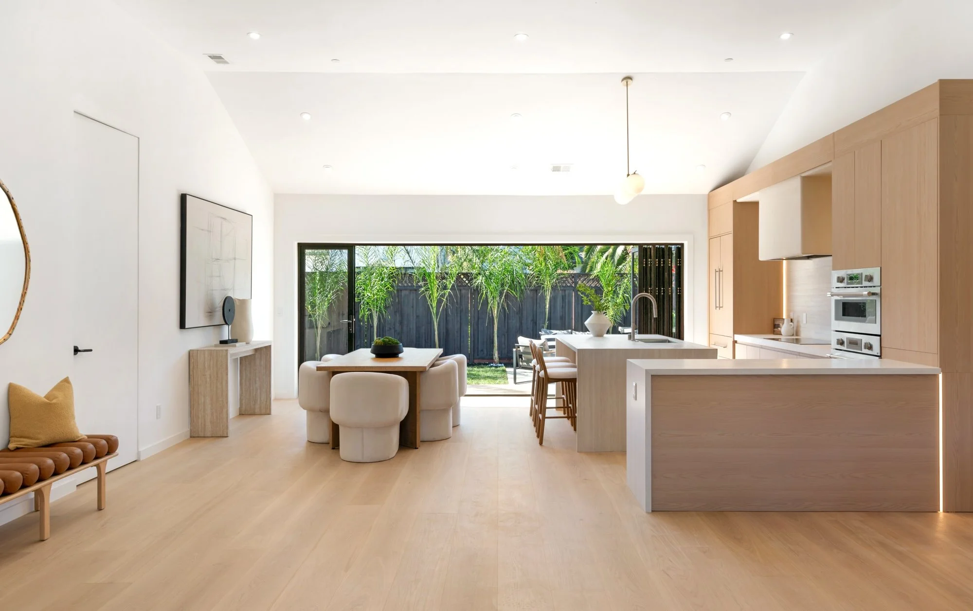 Open-concept modern kitchen and dining area with large sliding glass doors opening to a backyard with greenery, light wood flooring, white walls, and minimalistic decor.