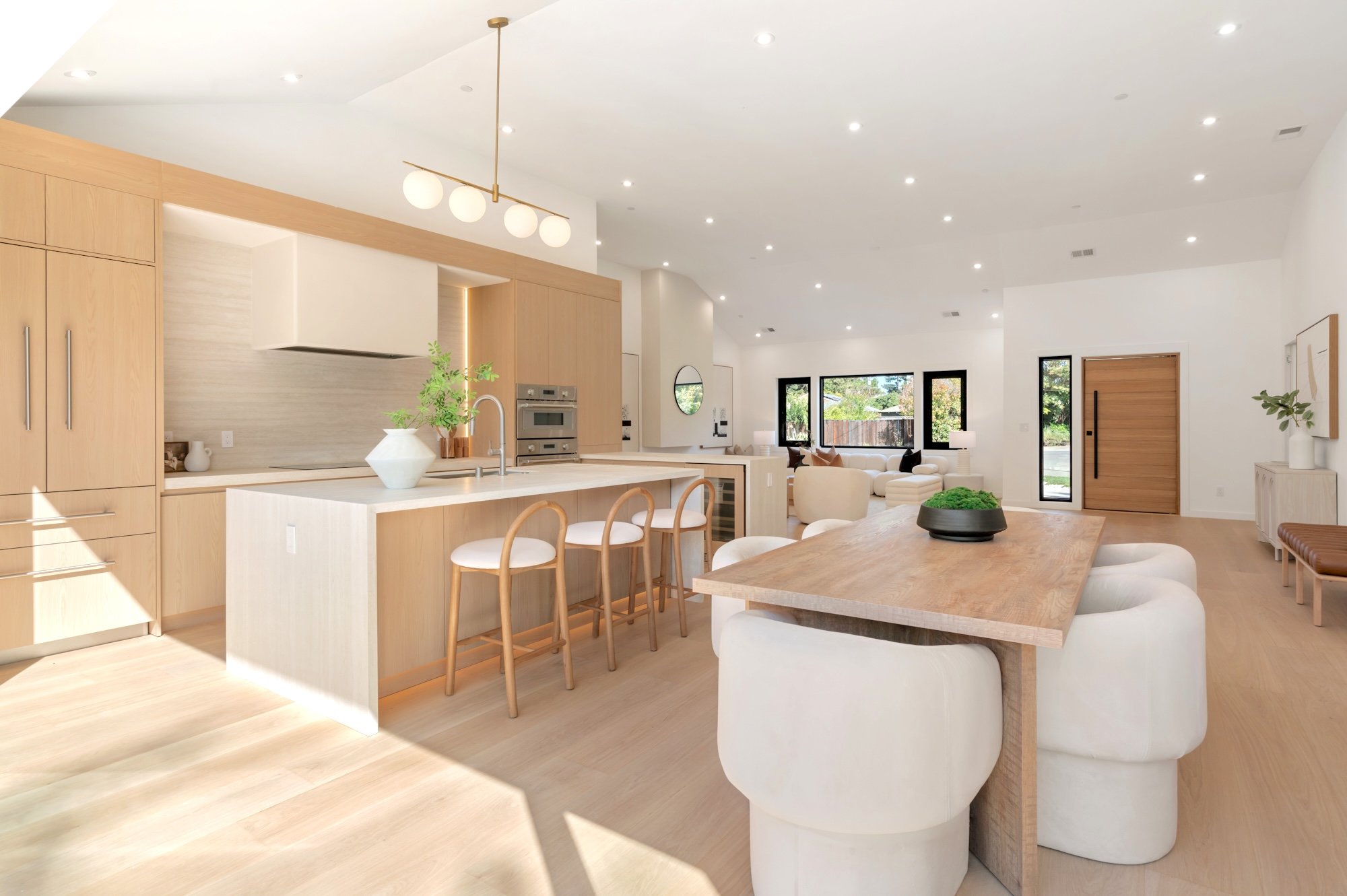 Open-concept living space with a kitchen and living area, featuring light wood cabinetry, white walls, a large wooden dining table, white chairs, and a neutral color palette with large windows letting in natural light.