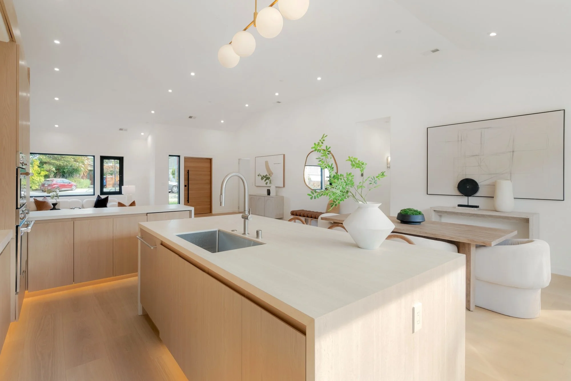 Modern open-concept kitchen and living room with white walls, wooden accents, and large windows, decorated with plants and minimalist artwork.