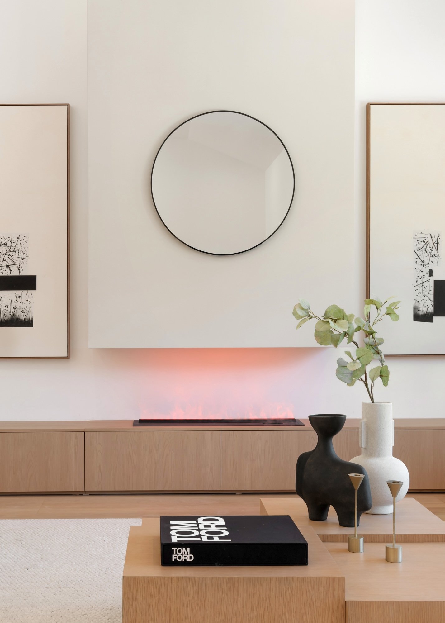 Minimalist living room with a white wall, circular mirror, wooden sideboard, abstract black and white artworks, a black and white Tom Ford book, and decorative vases with greenery.